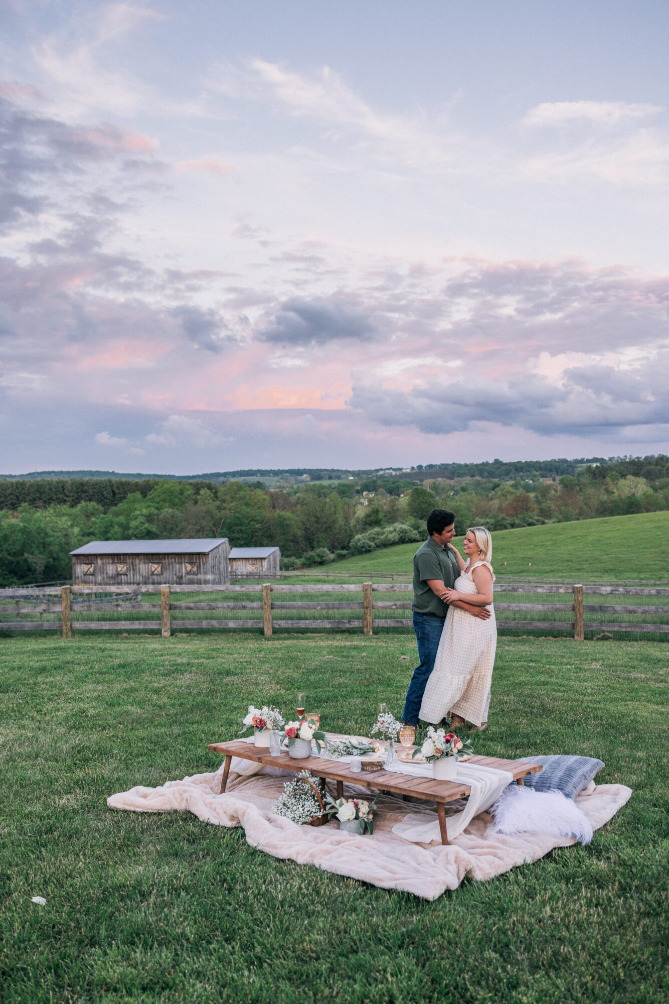 Two people standing close together on a grassy field, with a picnic setup and a fence in the background under a cloudy sky.