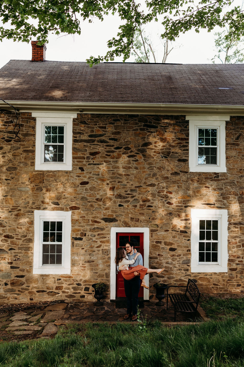 Person standing in front of a stone house with four windows and a red door, holding a guitar, outdoors.