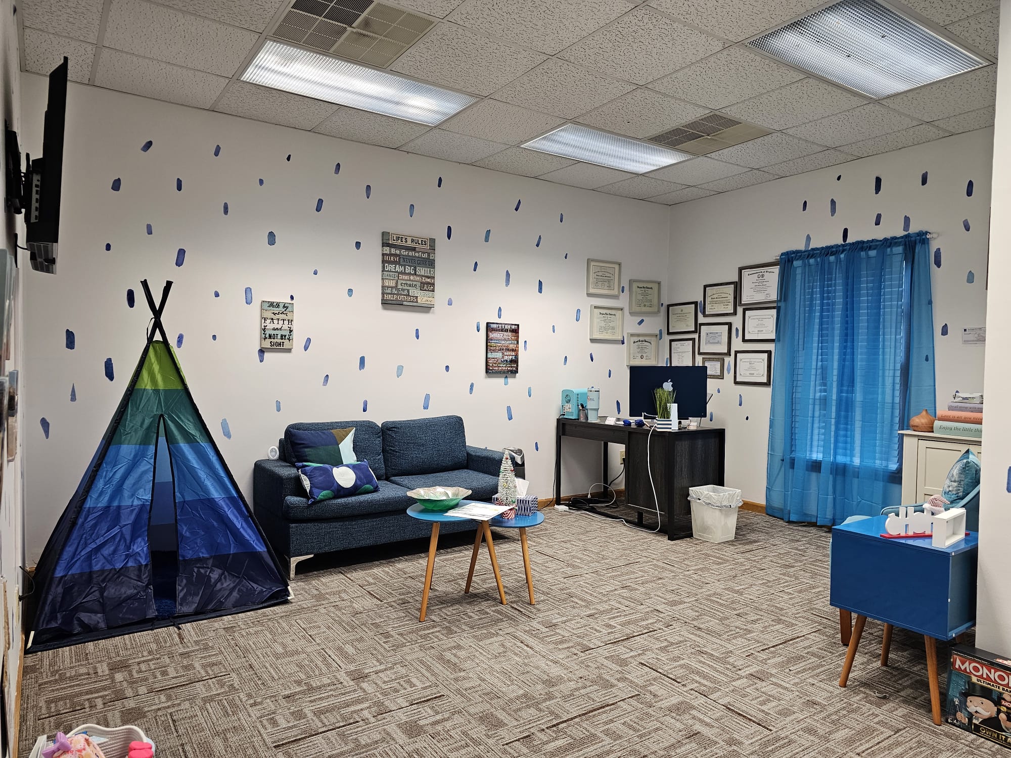 Indoor room with a blue tent, sofa, small table, computer desk, and blue curtains, decorated with framed pictures on the wall.