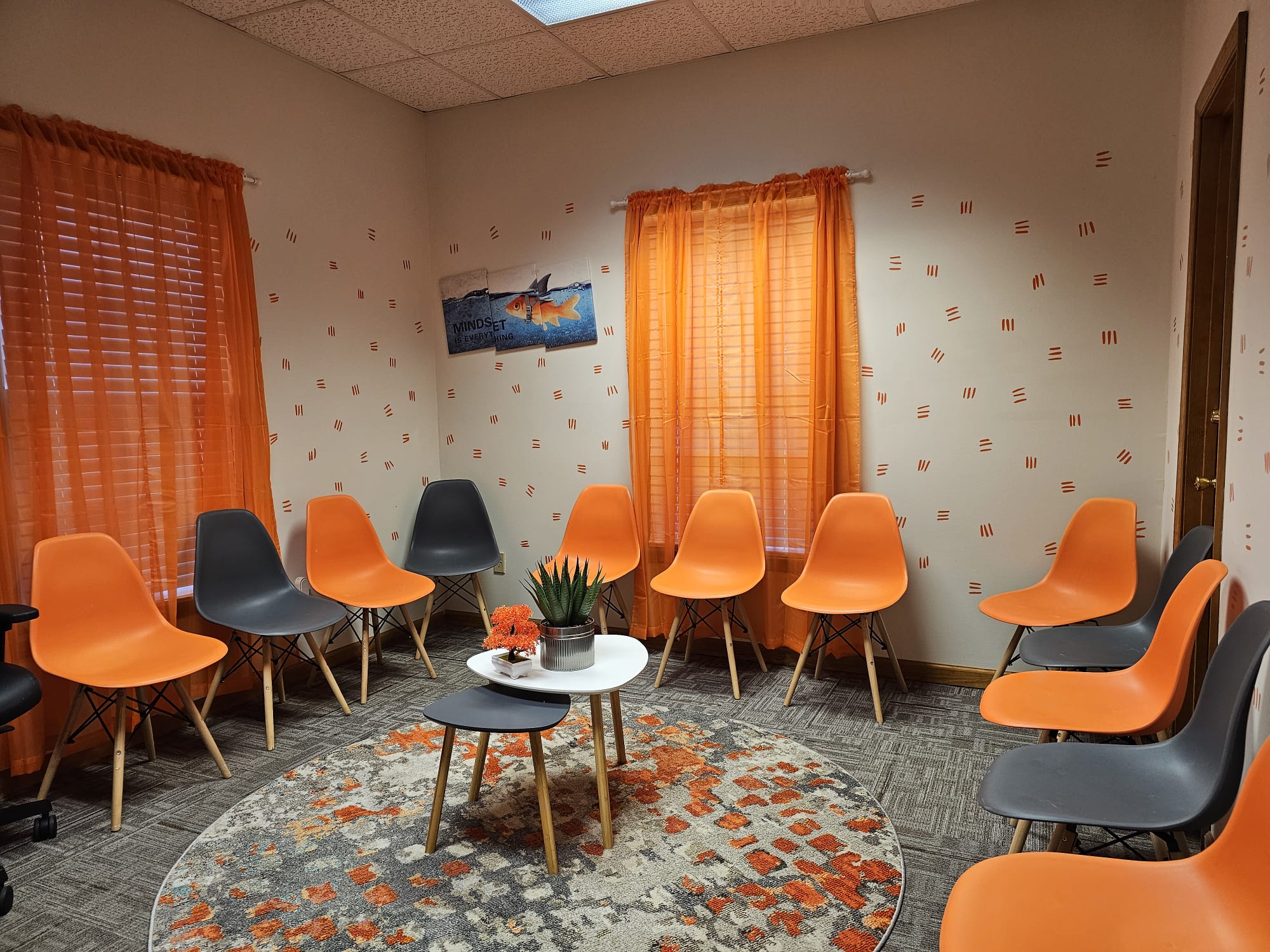 Room with orange and gray chairs arranged in a circle, two windows with orange curtains, a round rug, and a small table with a plant.