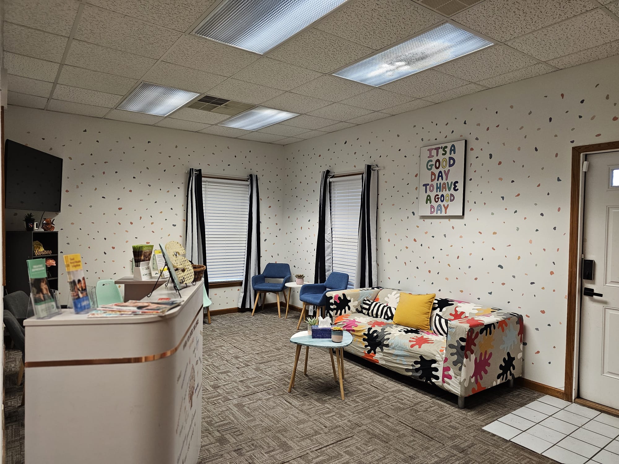 Waiting area with a floral sofa, two chairs, a small table, and a wall sign, in a room with polka dot wallpaper.