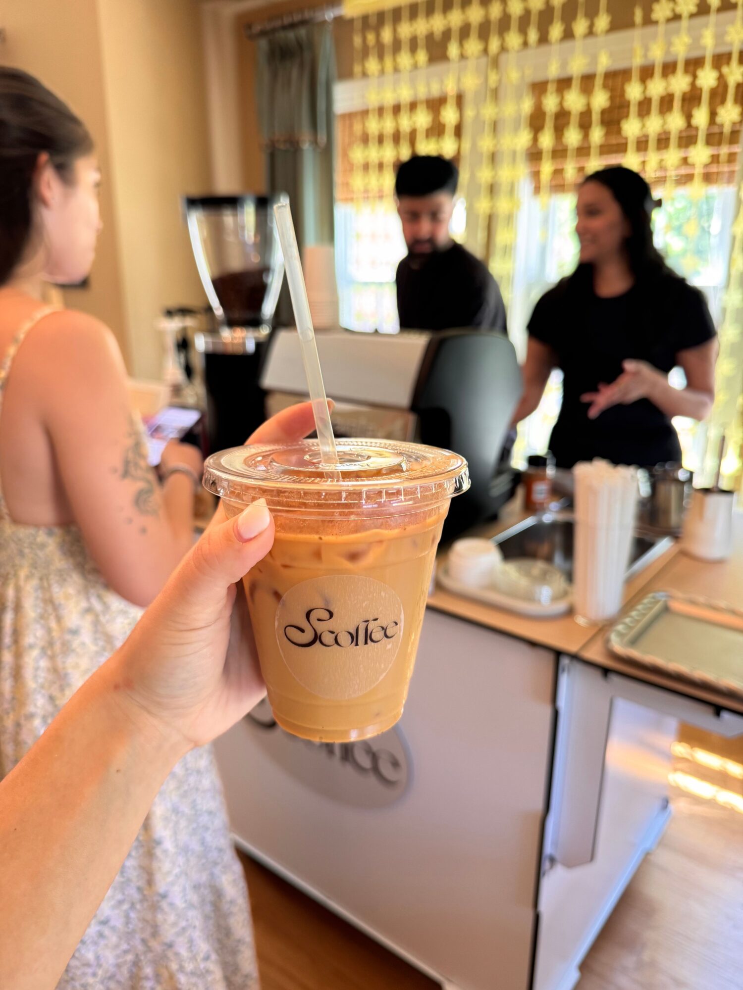 Hand holding iced coffee with straw in a cafe, with people in the background, natural light, and a window.