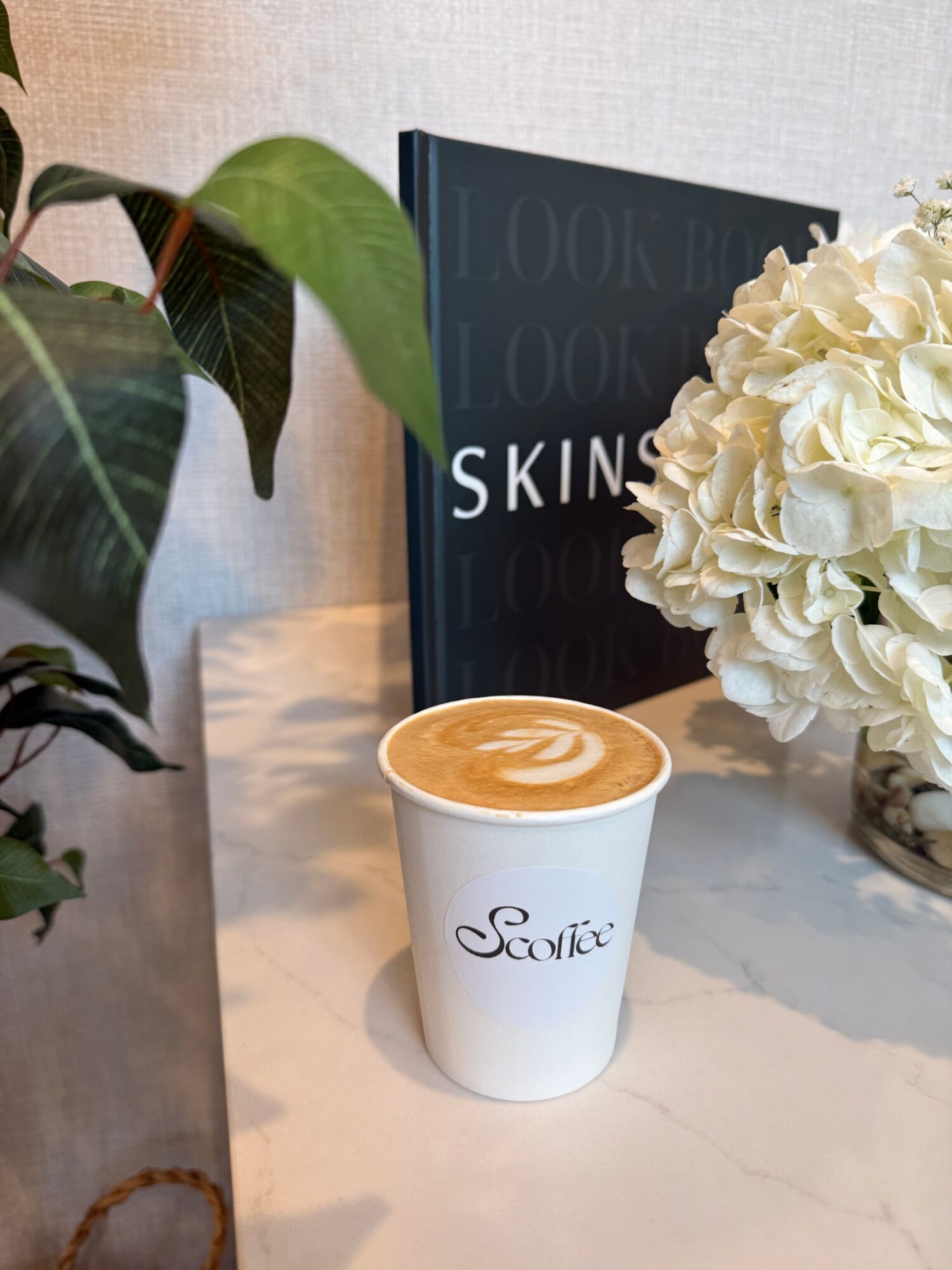 Coffee cup with latte art, white hydrangea flowers, a black book, and green leaves on a white marble surface.