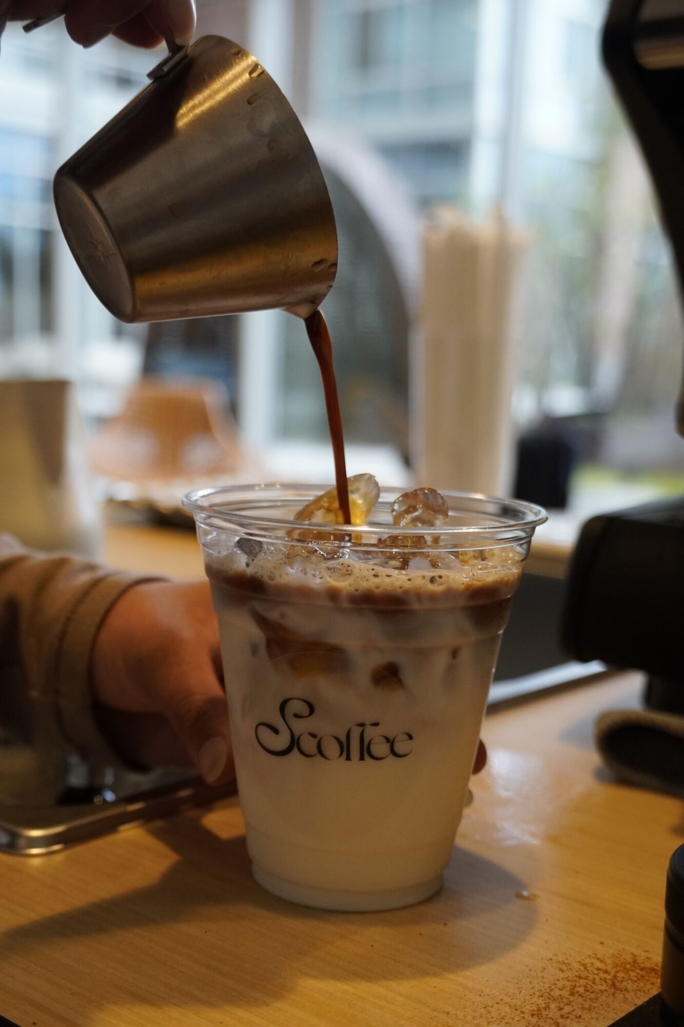Pouring coffee into a glass with ice and milk, on a wooden table in a cafe setting.