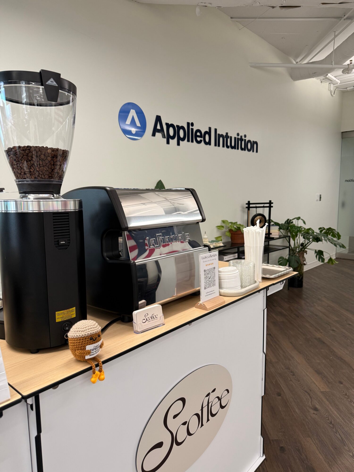 Office reception area with a counter, computer, and decorative plants, with a wall displaying the Applied Intuition logo.