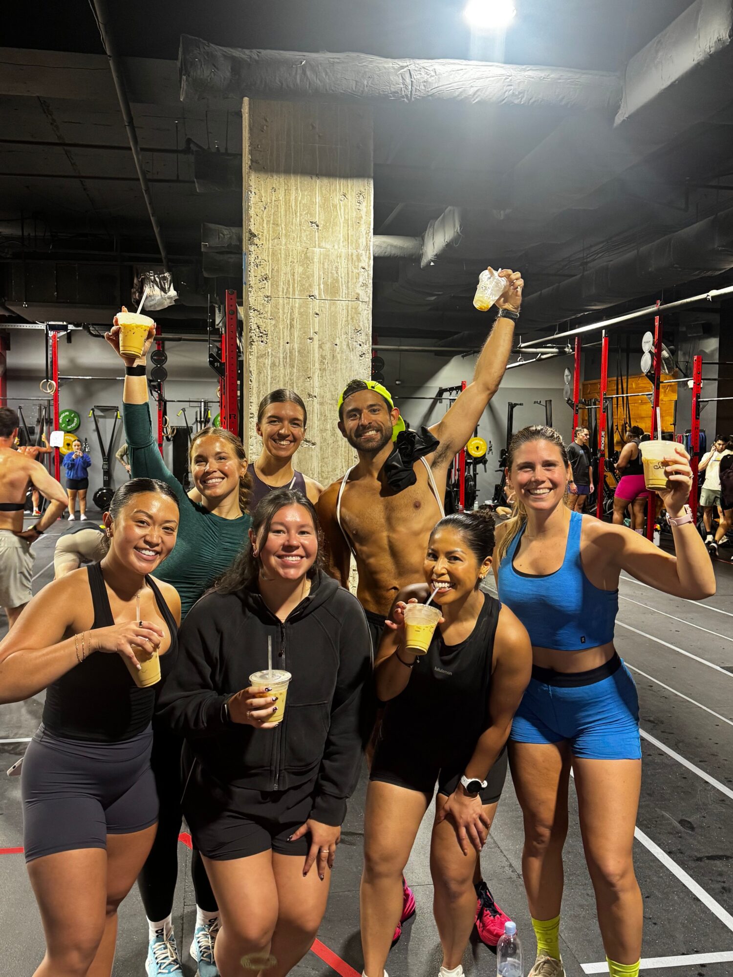Group of people celebrating with drinks in a gym or fitness studio, some wearing workout clothes, holding trophies, smiling.