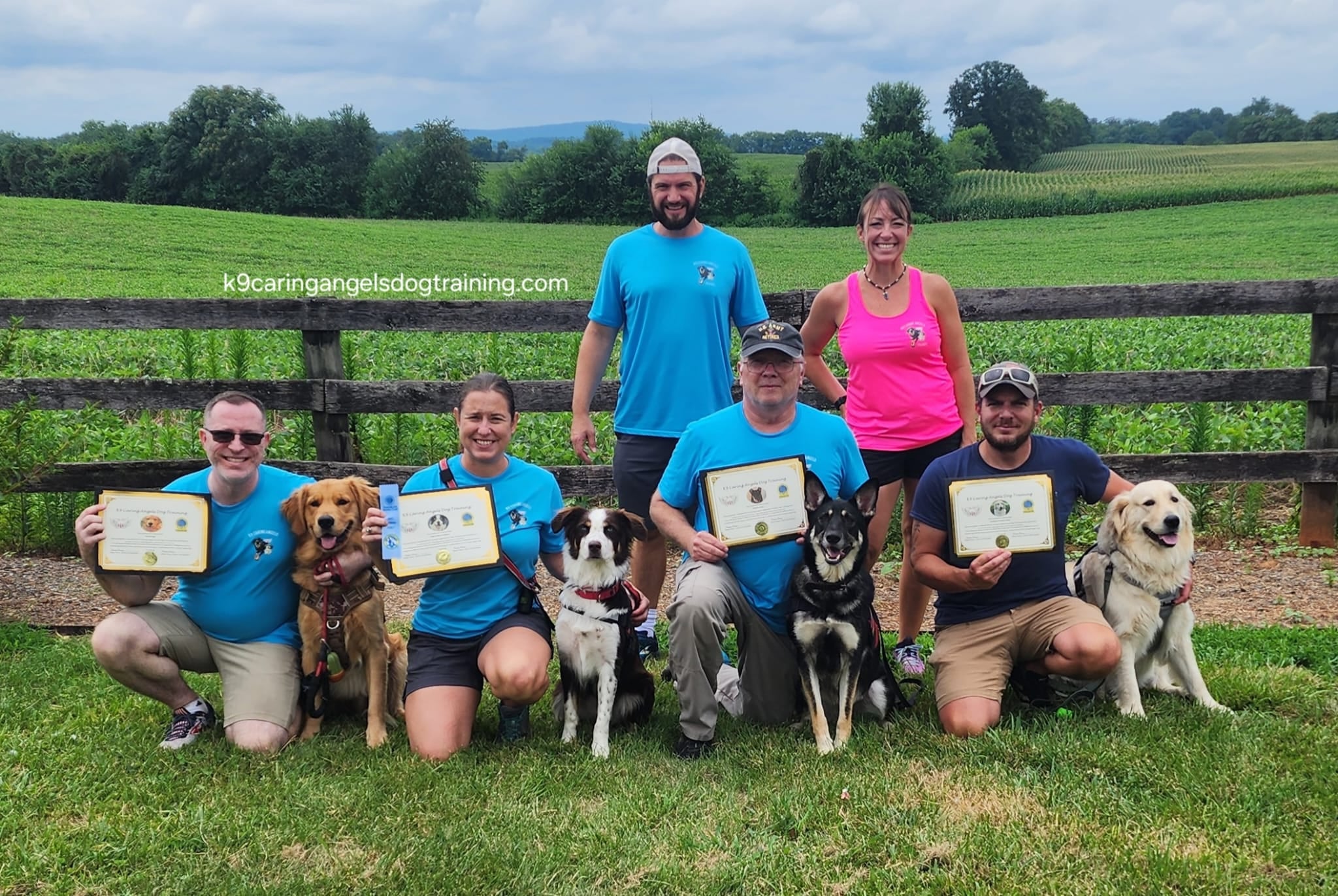Group of six people and five dogs outdoors on grass, holding certificates, with a wooden fence and green fields in background.