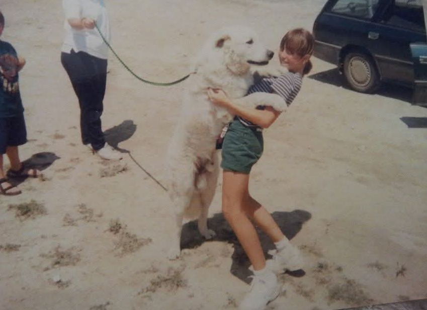 Child hugging a large dog outdoors with other people and a parked car nearby.