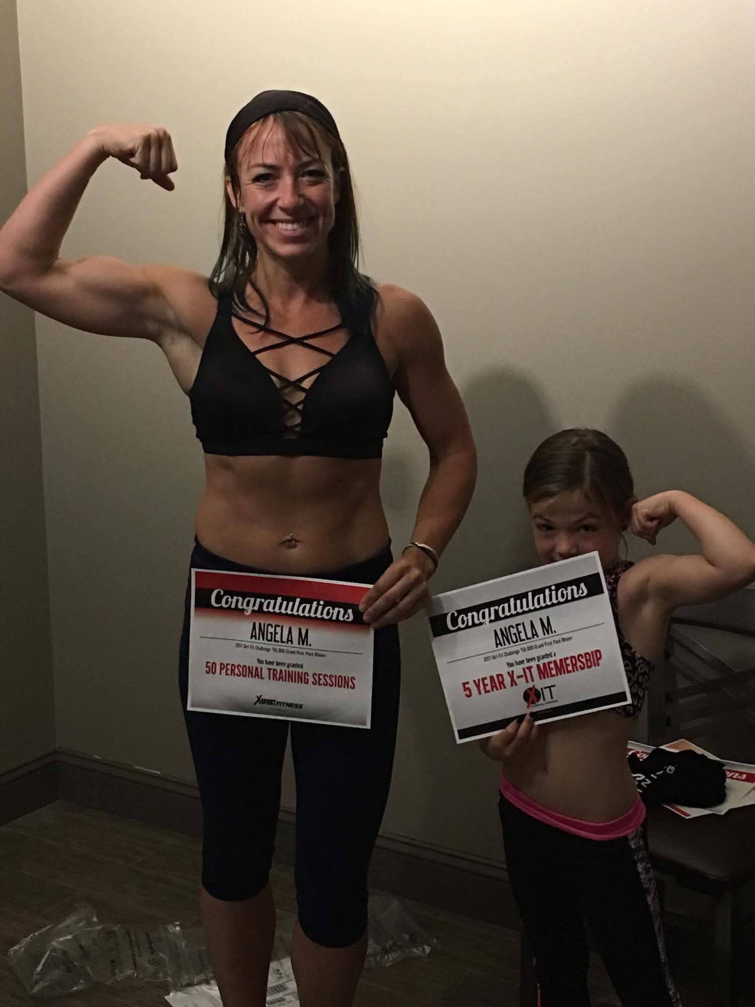 Woman and girl flexing muscles, holding congratulatory signs, standing indoors against a plain wall.