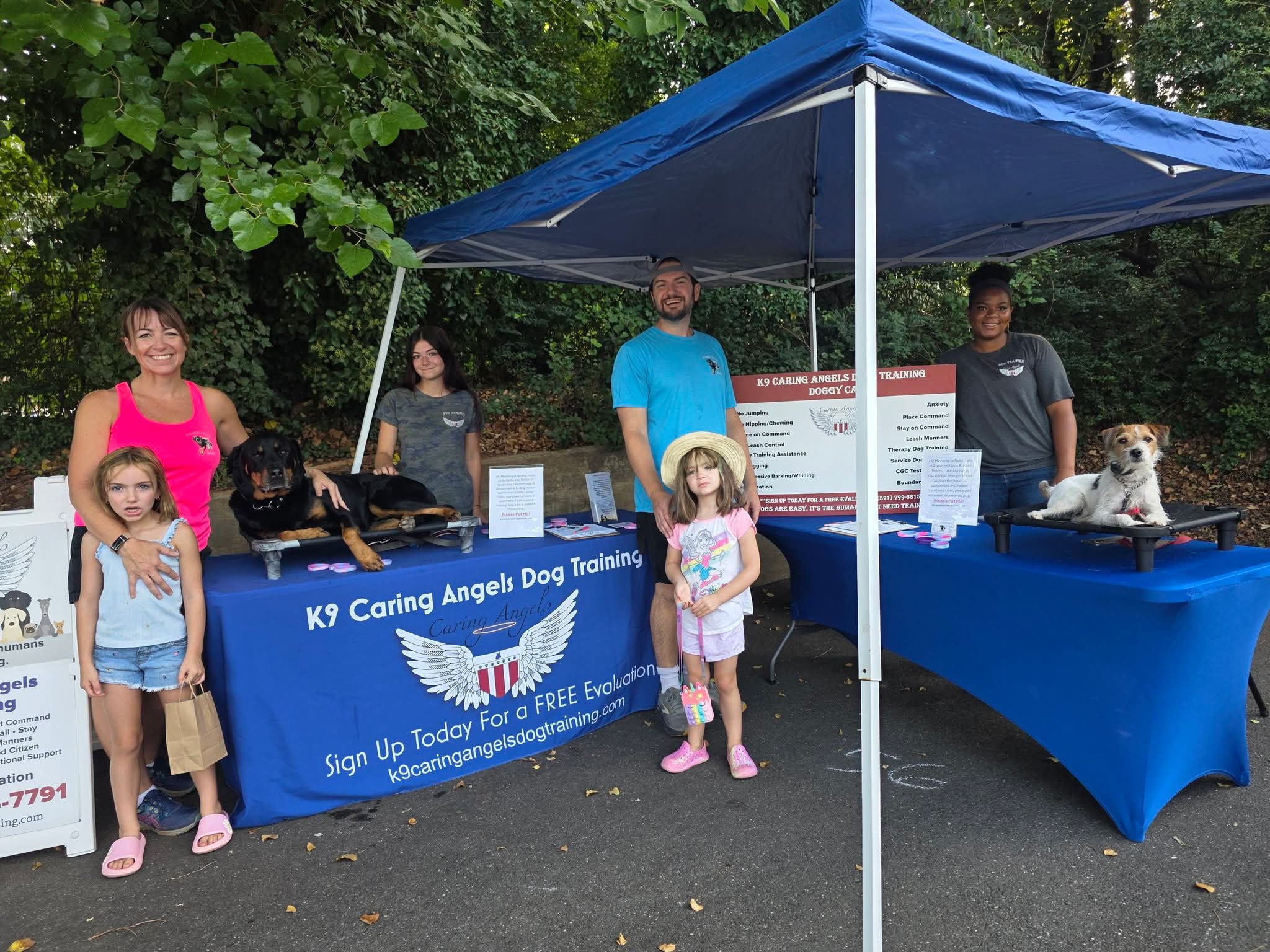 Group of children and adults at a dog adoption event with two tables and dogs, outdoors with trees in background.