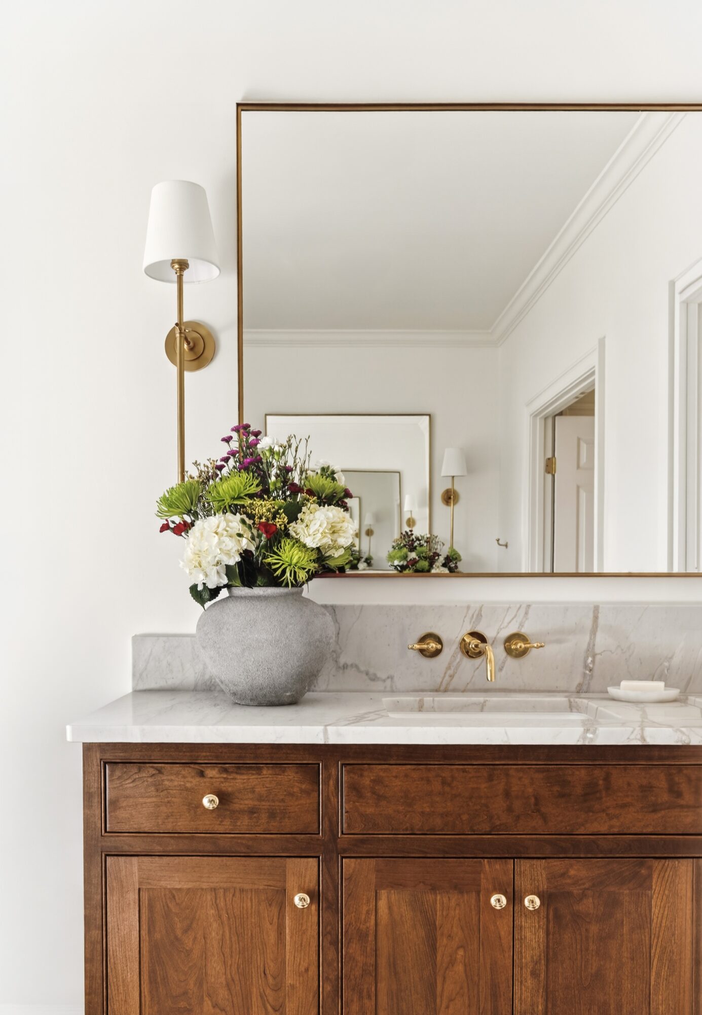 Bathroom vanity with a large mirror, flower arrangement, and wall-mounted light fixture, in a bright, white room.