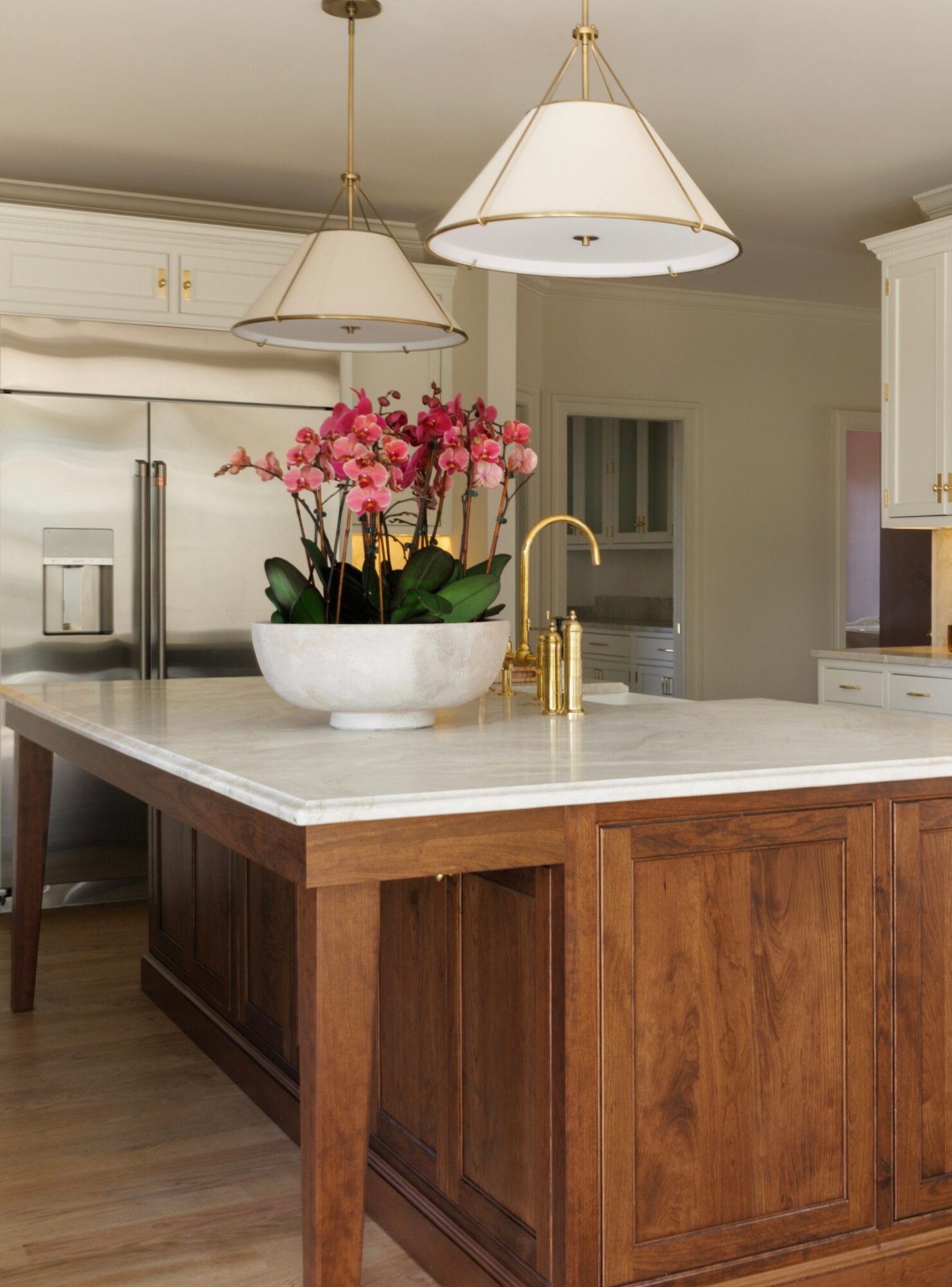 Kitchen island with a white marble top, wooden base, and a white bowl with pink flowers, gold faucet, and pendant lights above.