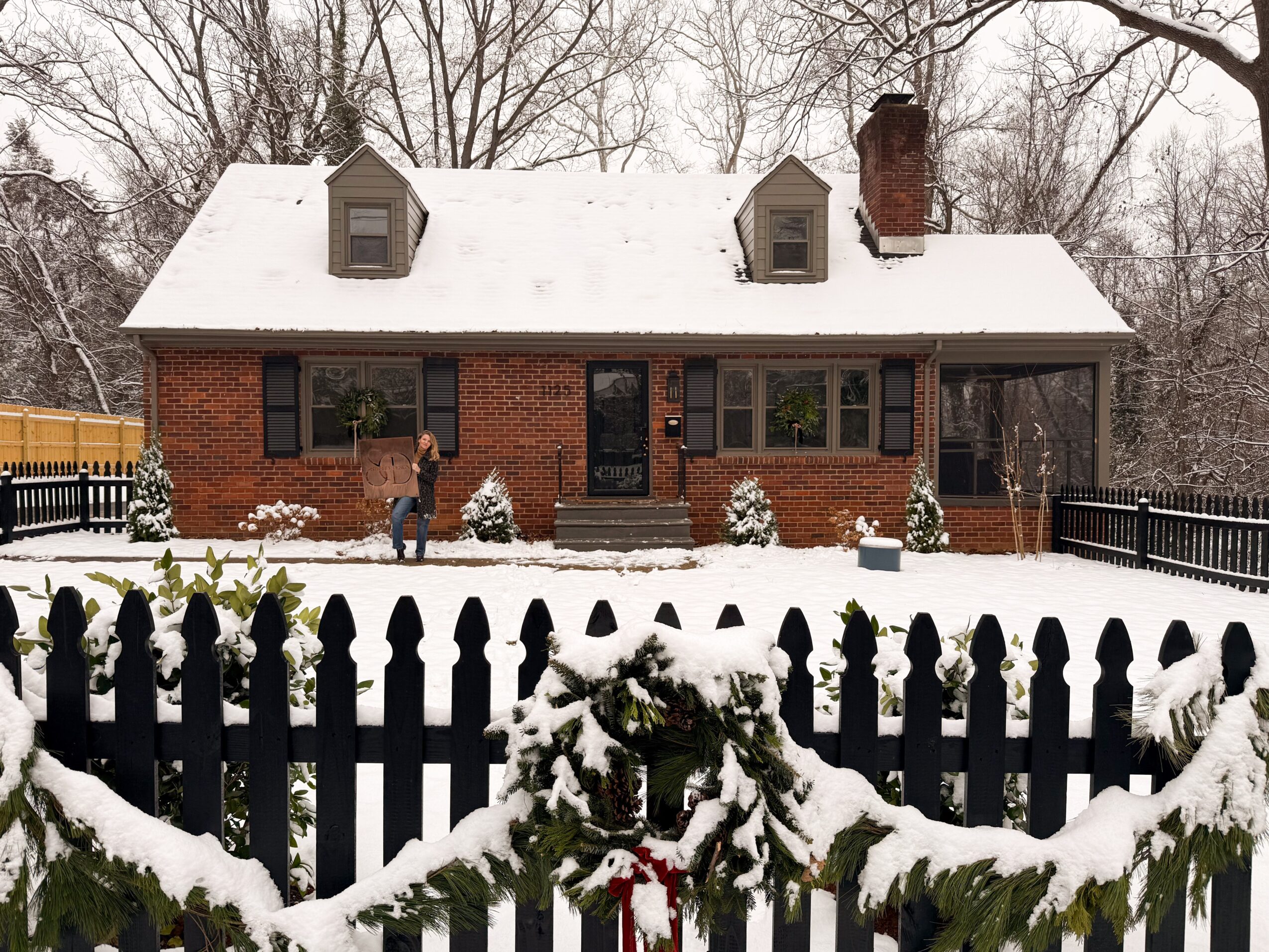 Brick house with snow-covered roof and yard, black fence with snow, trees in background, two people near house, winter scene.