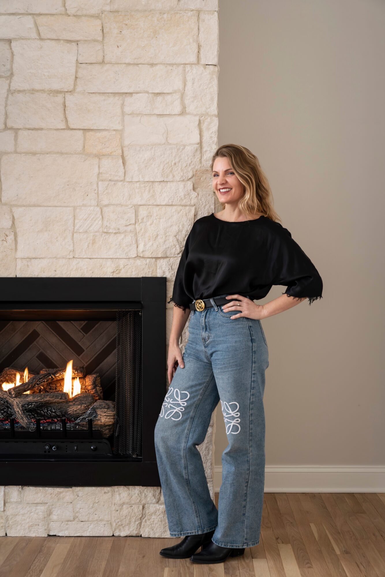 Woman standing next to a fireplace with a stone wall, smiling, wearing a black top and jeans, hands on hips.
