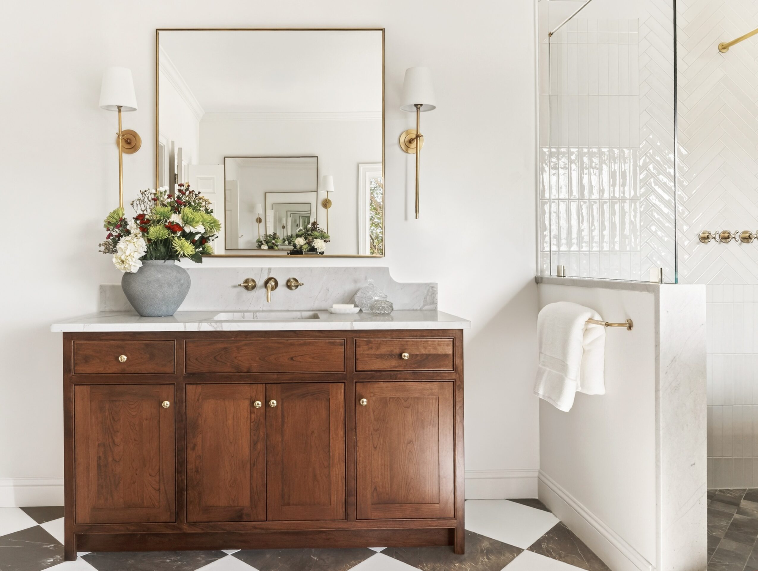 Bathroom vanity with a large mirror, flower arrangement, and wall-mounted lights, with a towel on a nearby rack.