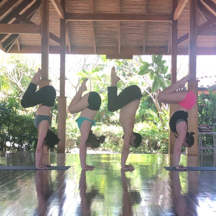 Four people practicing yoga in a wooden pavilion with greenery outside, performing handstands and forward bends.