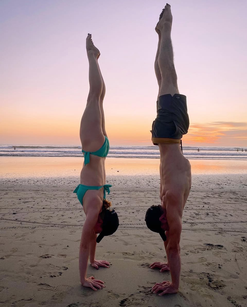 A man and woman perform handstands on the beach at sunset, facing each other with the ocean in the background.