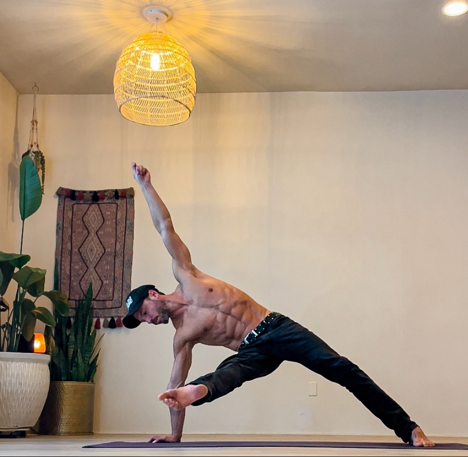 Person practicing yoga in a room with plants, a wall hanging, and a hanging light fixture, performing a side plank pose.
