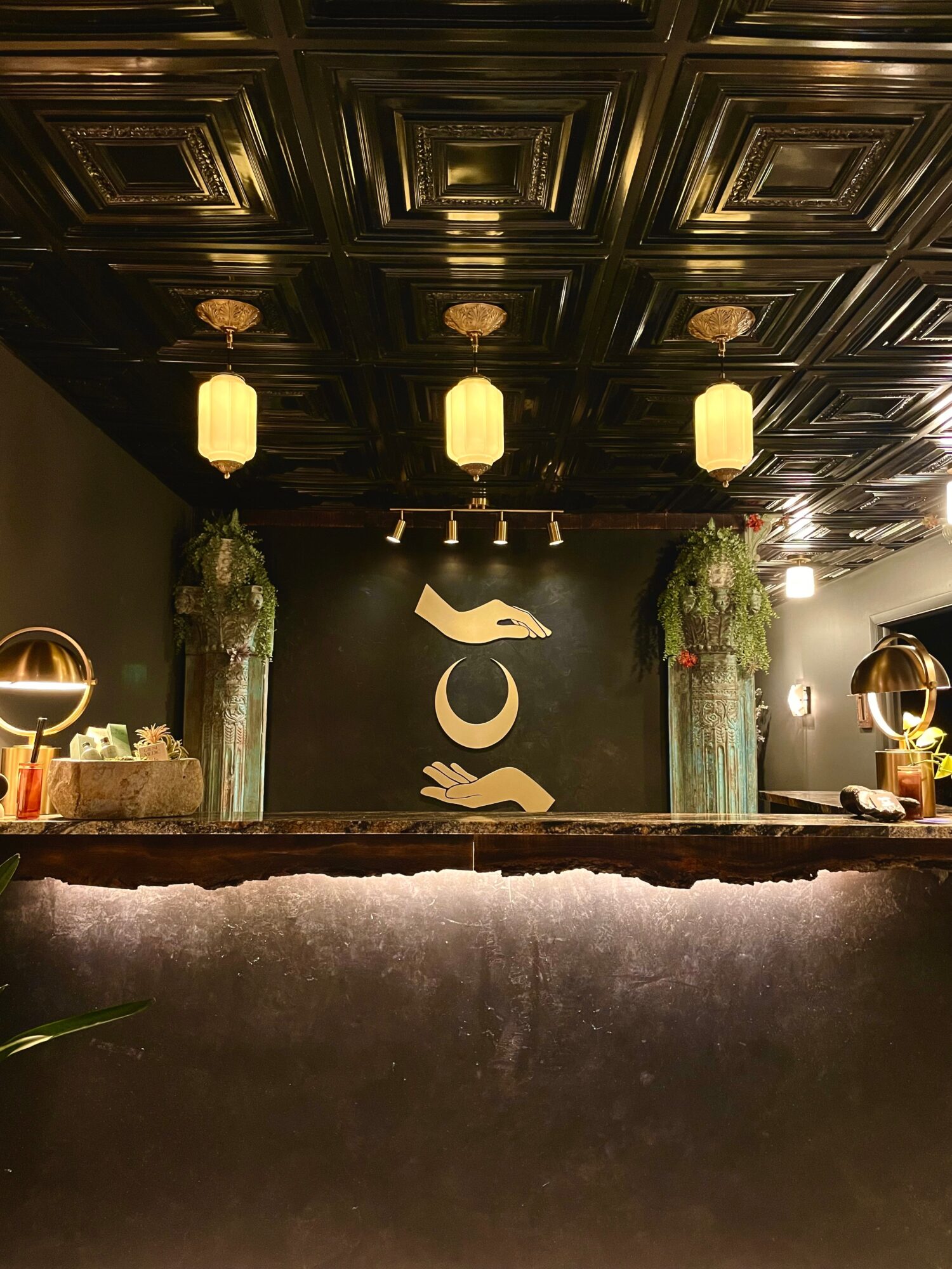 Elegant reception area with black wall, decorative symbols, two tall vases with flowers, and hanging pendant lights.