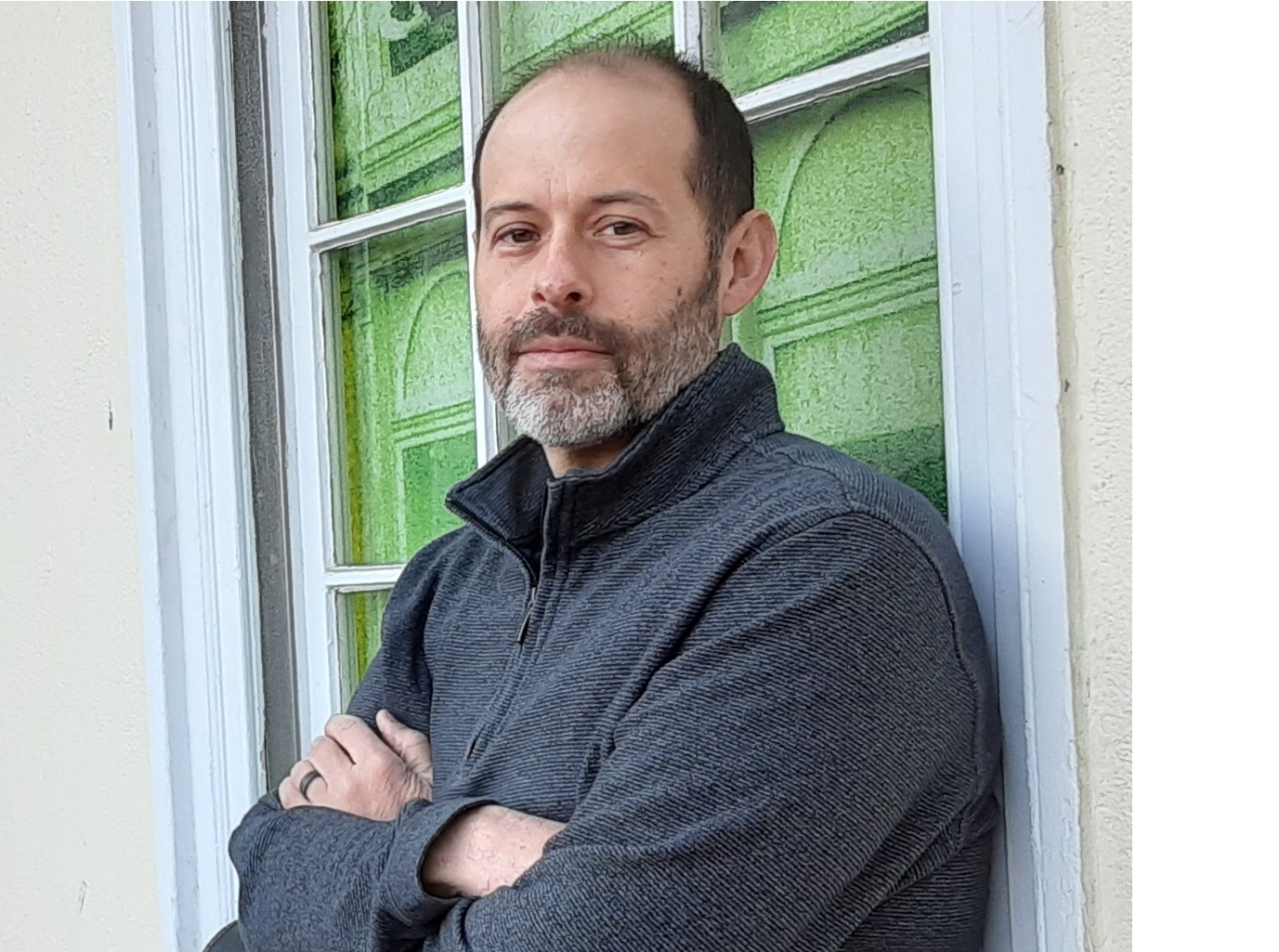 Man with a beard and short hair, wearing a dark zip-up jacket, standing outside near a window with green panels.