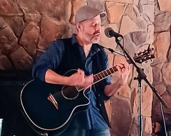 Man playing guitar and singing into microphone against a stone wall background.