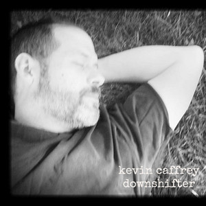 Man with short hair and beard lying on grass with eyes closed, wearing a dark shirt, relaxed arm behind head, black and white photo.