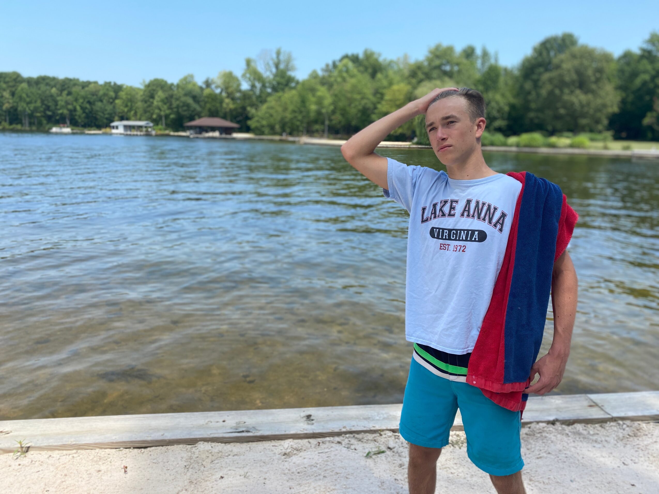 Young man standing by a lake with trees in the background, wearing a white T-shirt and blue shorts, holding a towel over his shoulder.