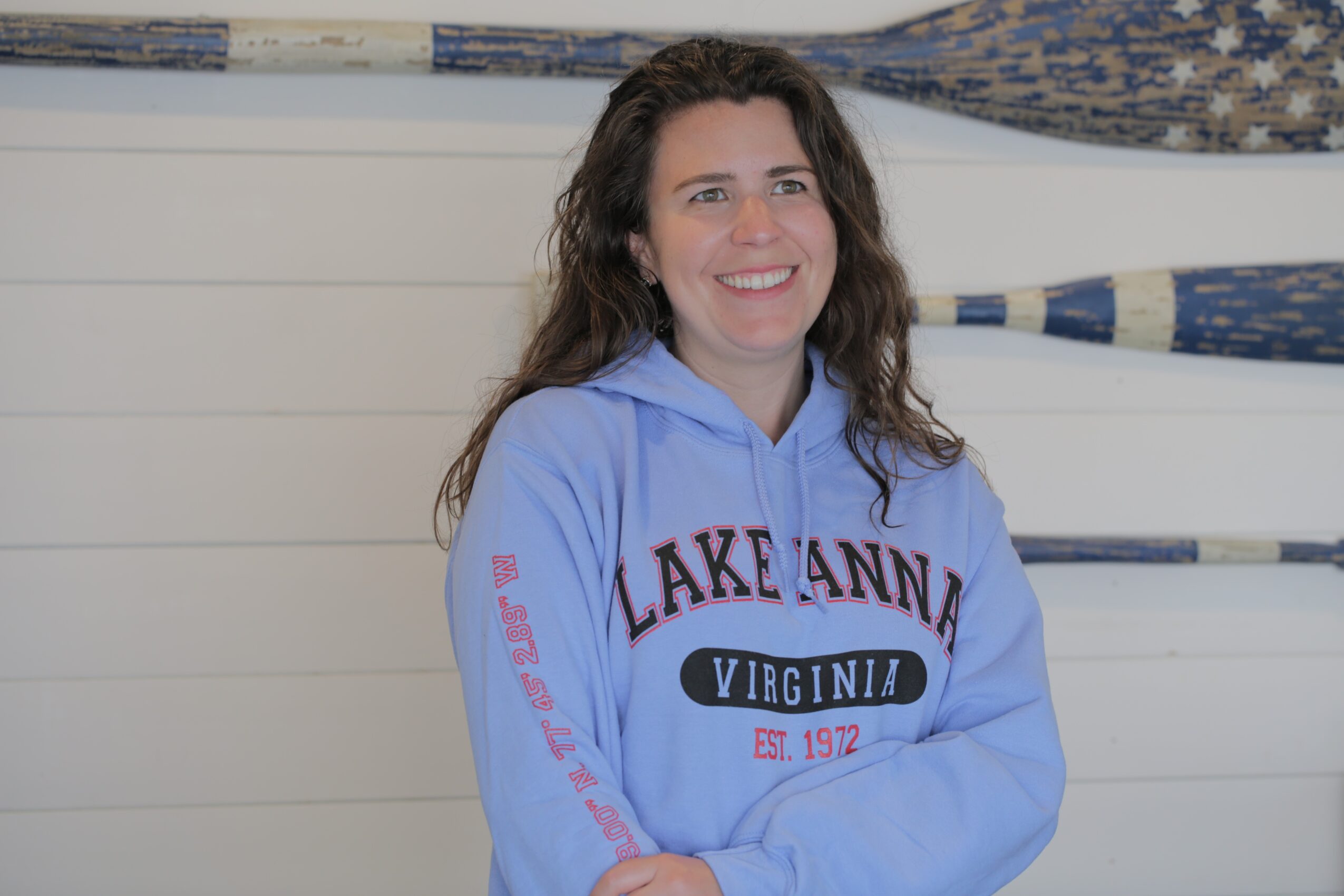Woman with long dark hair smiling, wearing a blue hoodie with text, standing in front of a white wall with paddles.