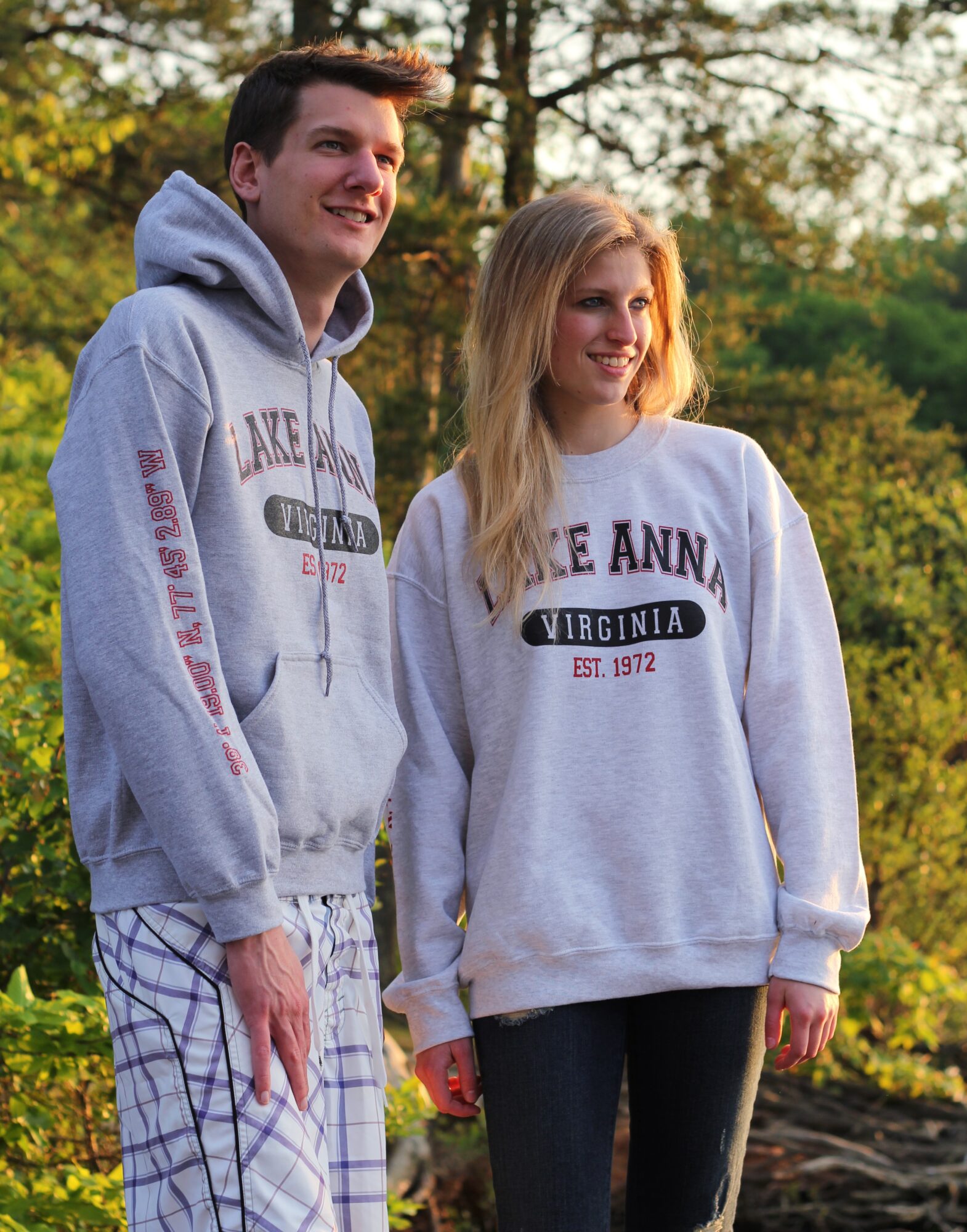 A young man and woman stand outdoors in a park with trees and sunlight, both wearing gray sweatshirts with Virginia text.