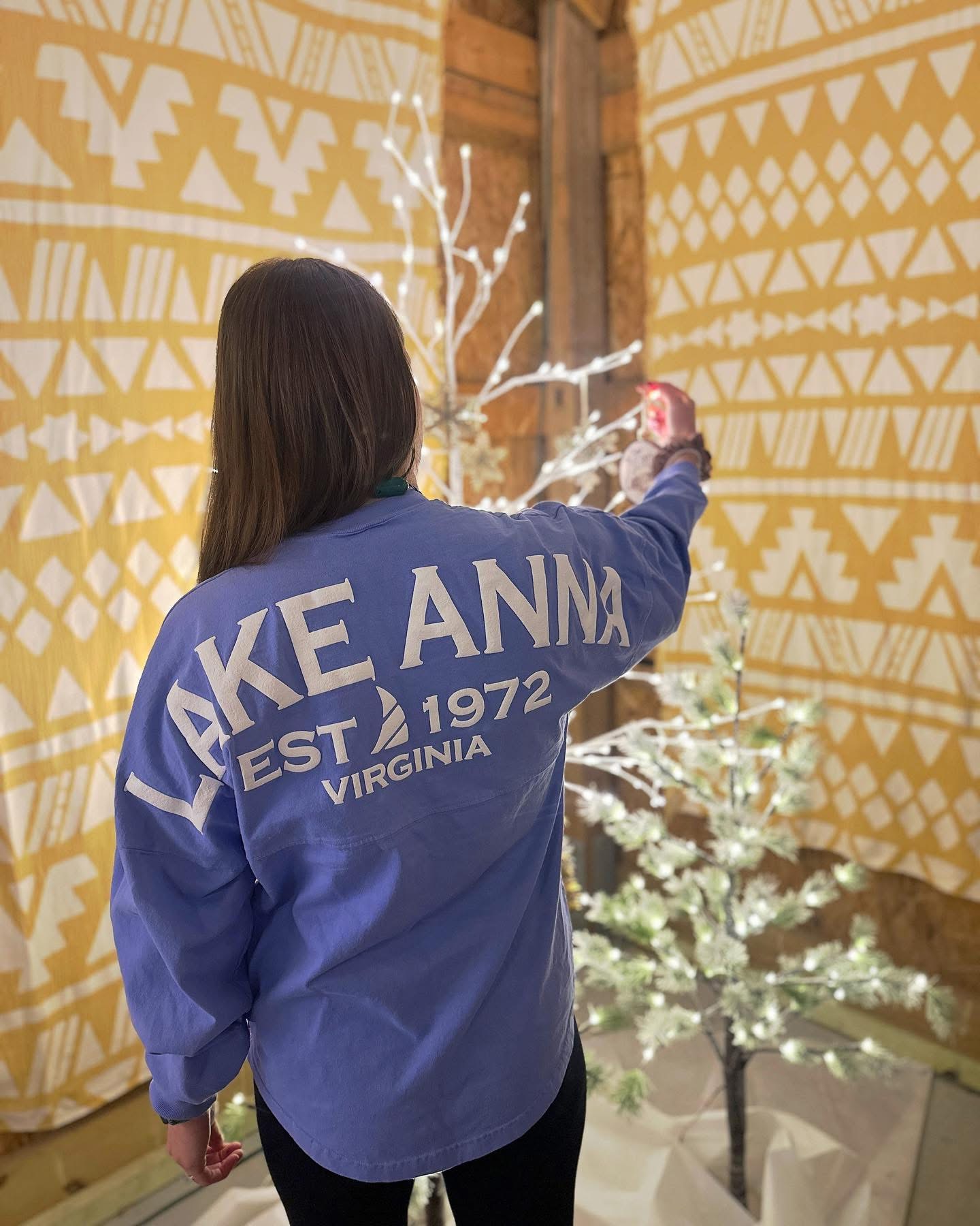 Person with long hair wearing a blue jacket with text, decorating a small Christmas tree with lights, indoors.