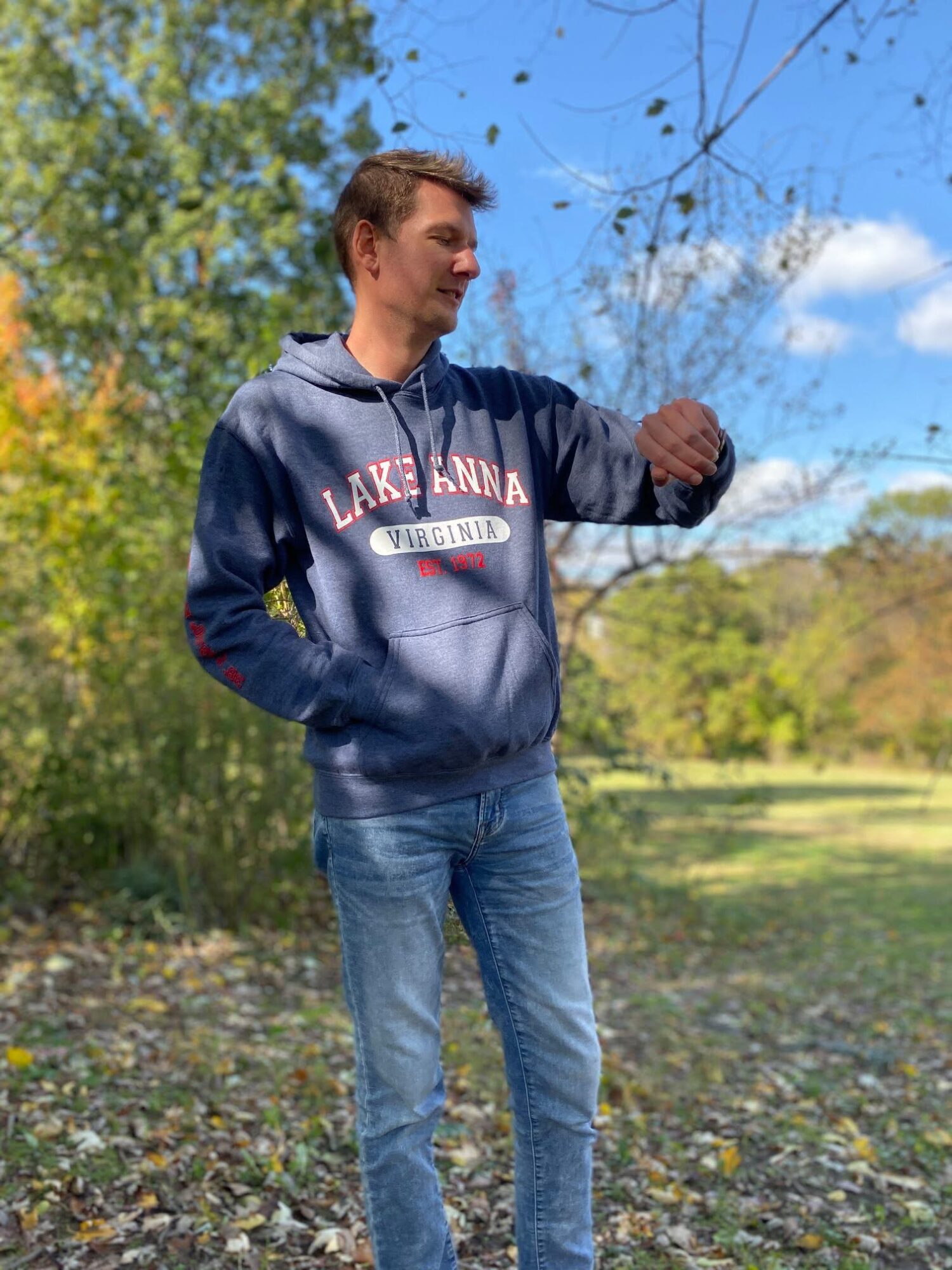 Young man standing outdoors in a park, looking at his watch, with trees and blue sky in the background.