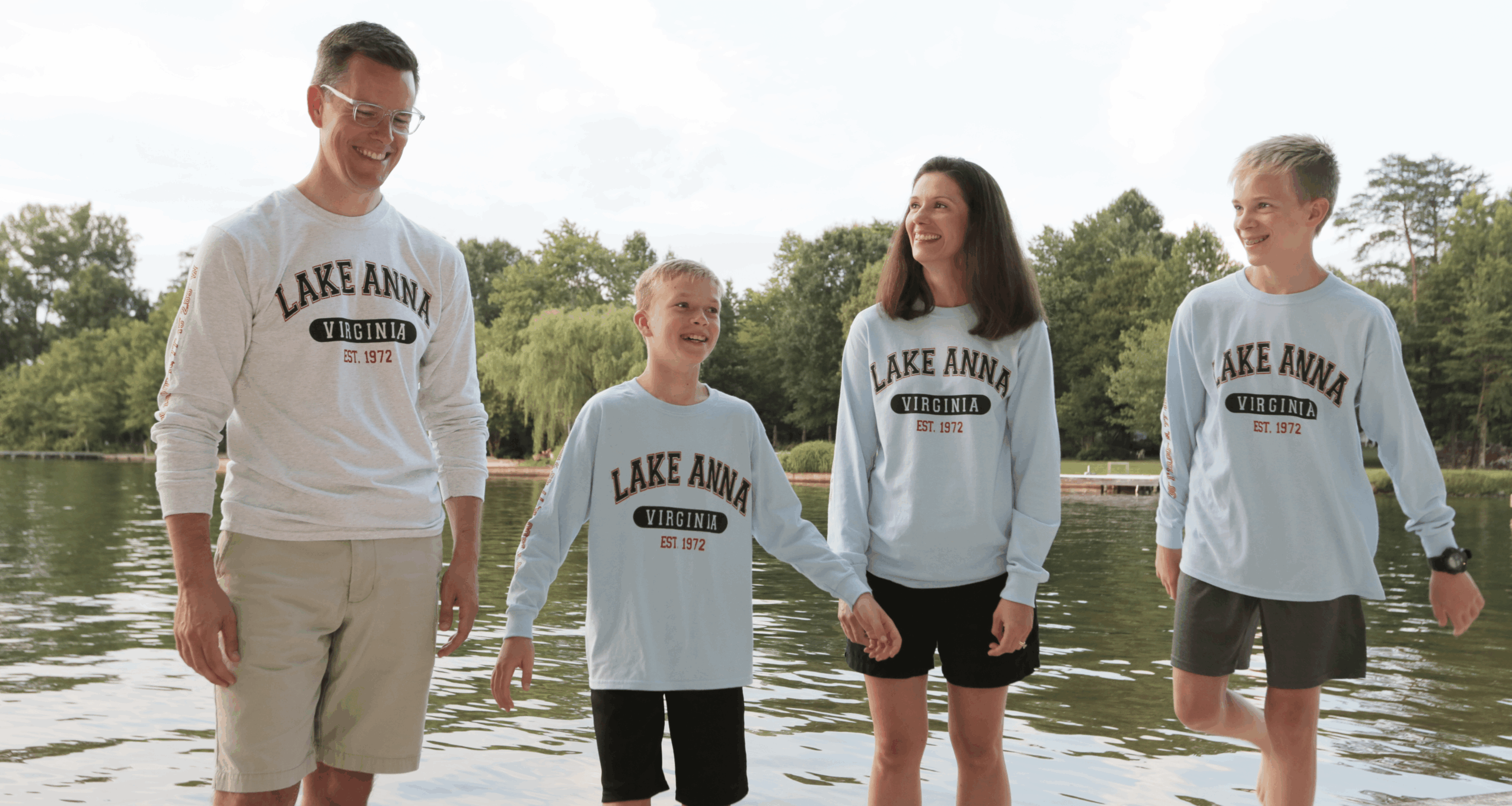 Four people standing by a lake, smiling and talking, with trees in the background.