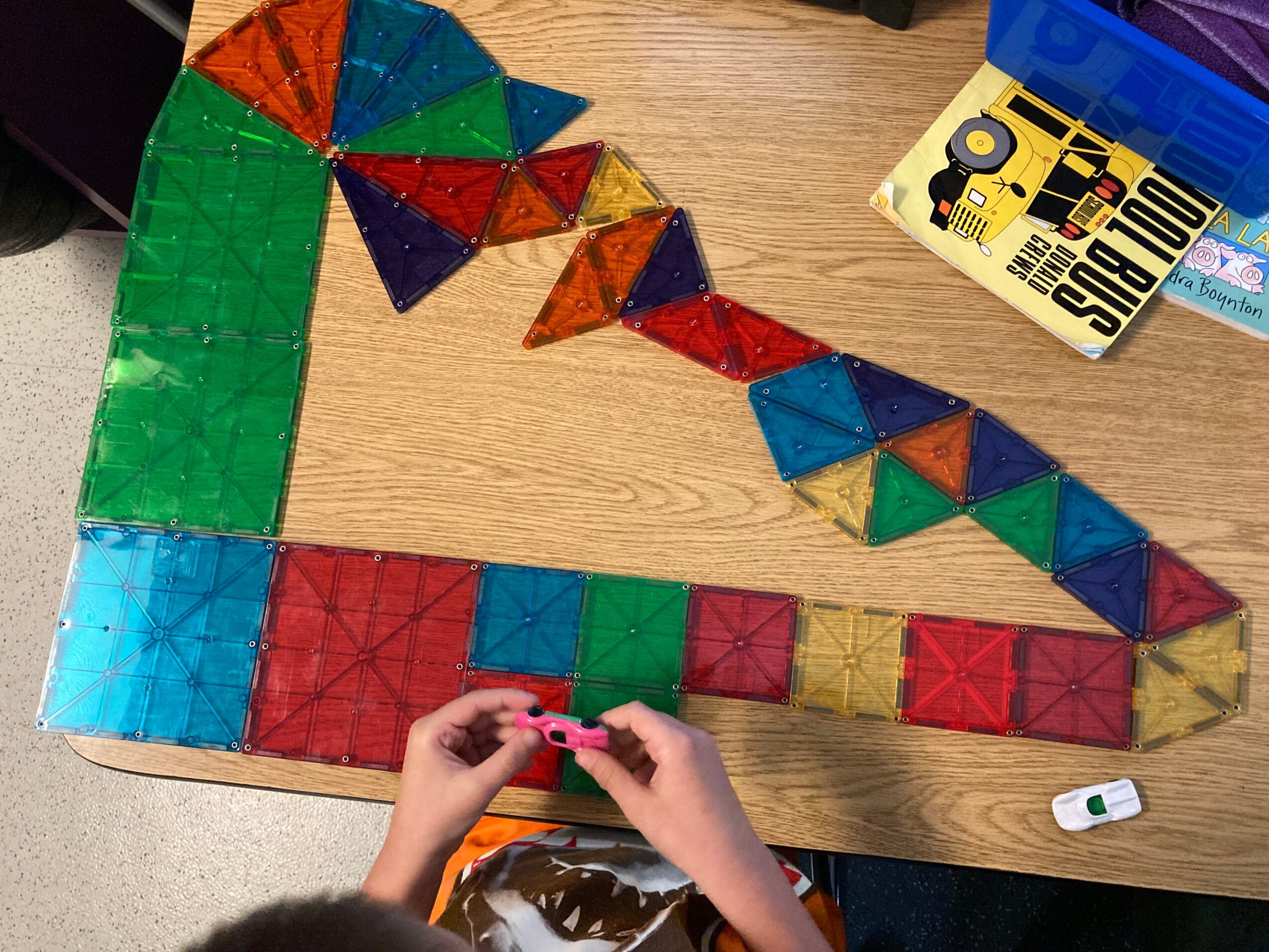 Colorful geometric shapes arranged on a wooden table, with a person's hands holding a small object, and a yellow box nearby.