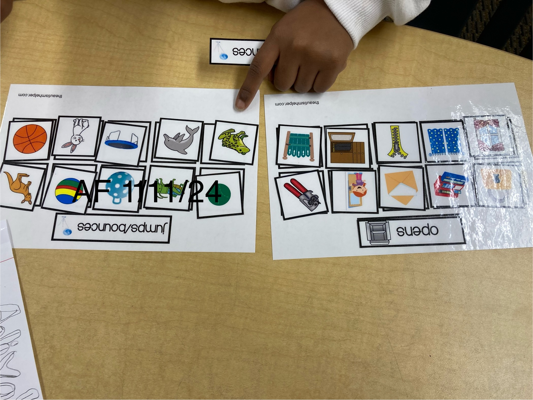 Two sheets of paper with pictures and words, a child's hand pointing at the left sheet, on a wooden table.