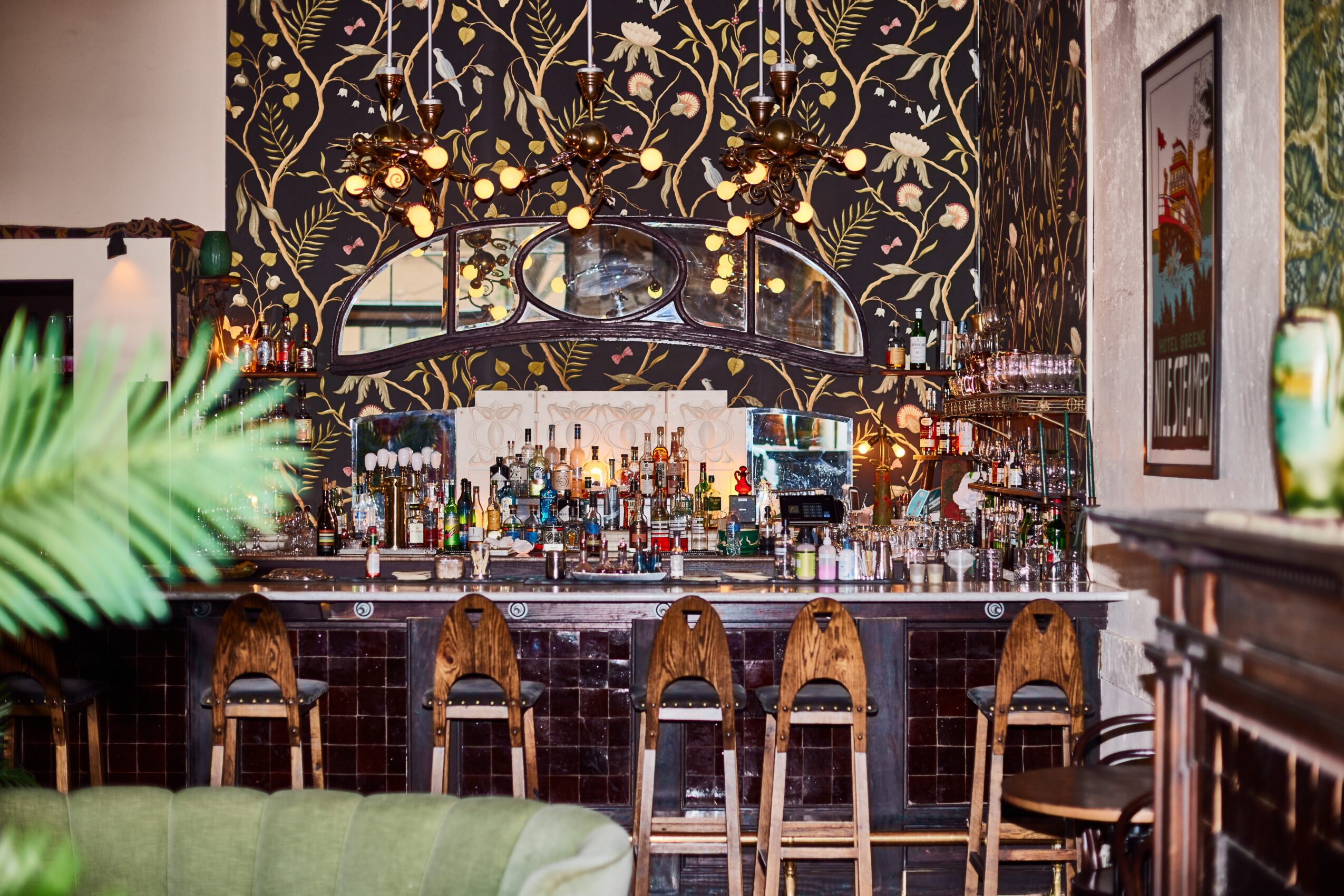Bar with a dark floral wallpaper background, illuminated hanging lights, and a variety of bottles behind the counter, with bar stools in front.
