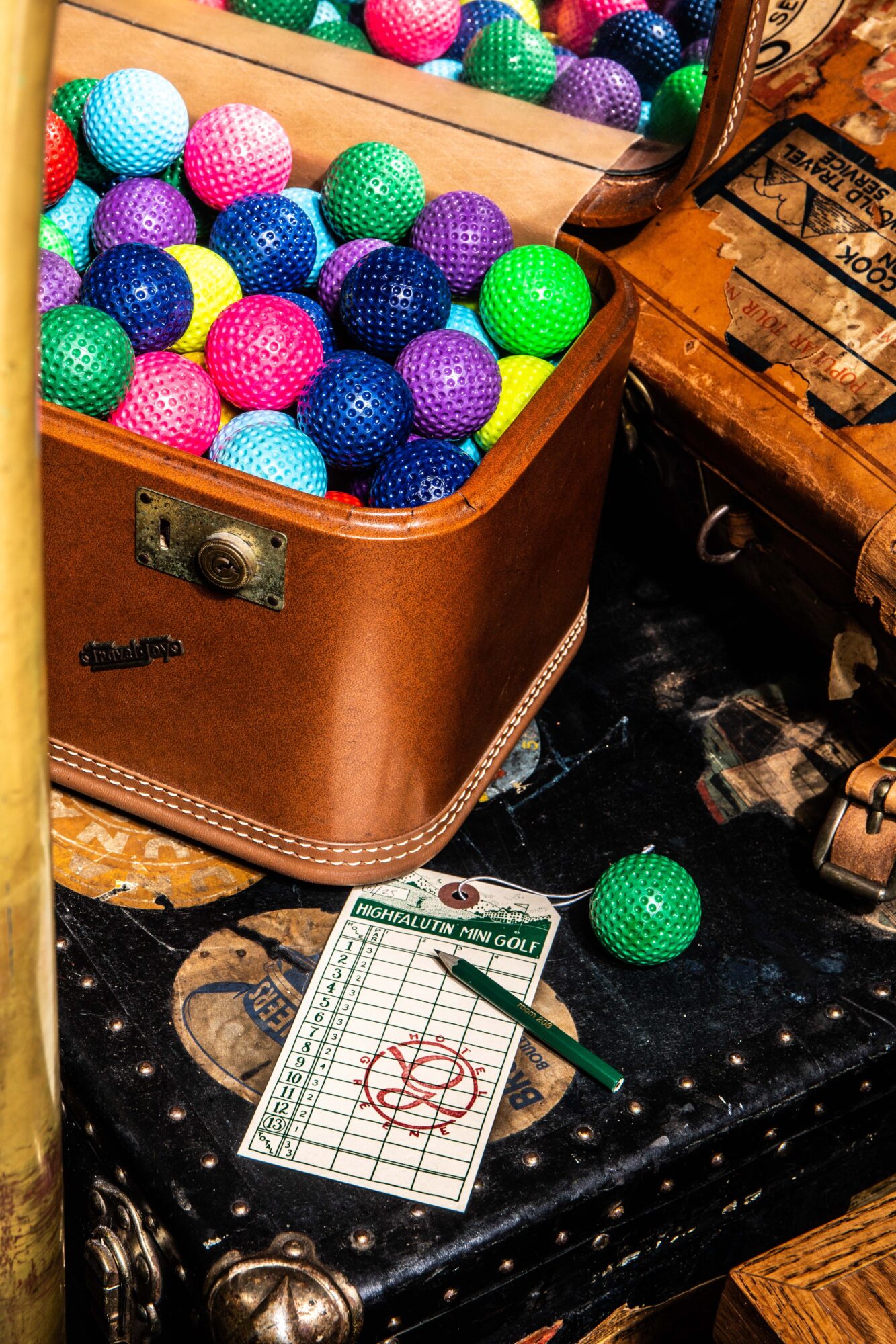 Open brown suitcase filled with colorful plastic balls, with a green ball and a score sheet on a black surface.