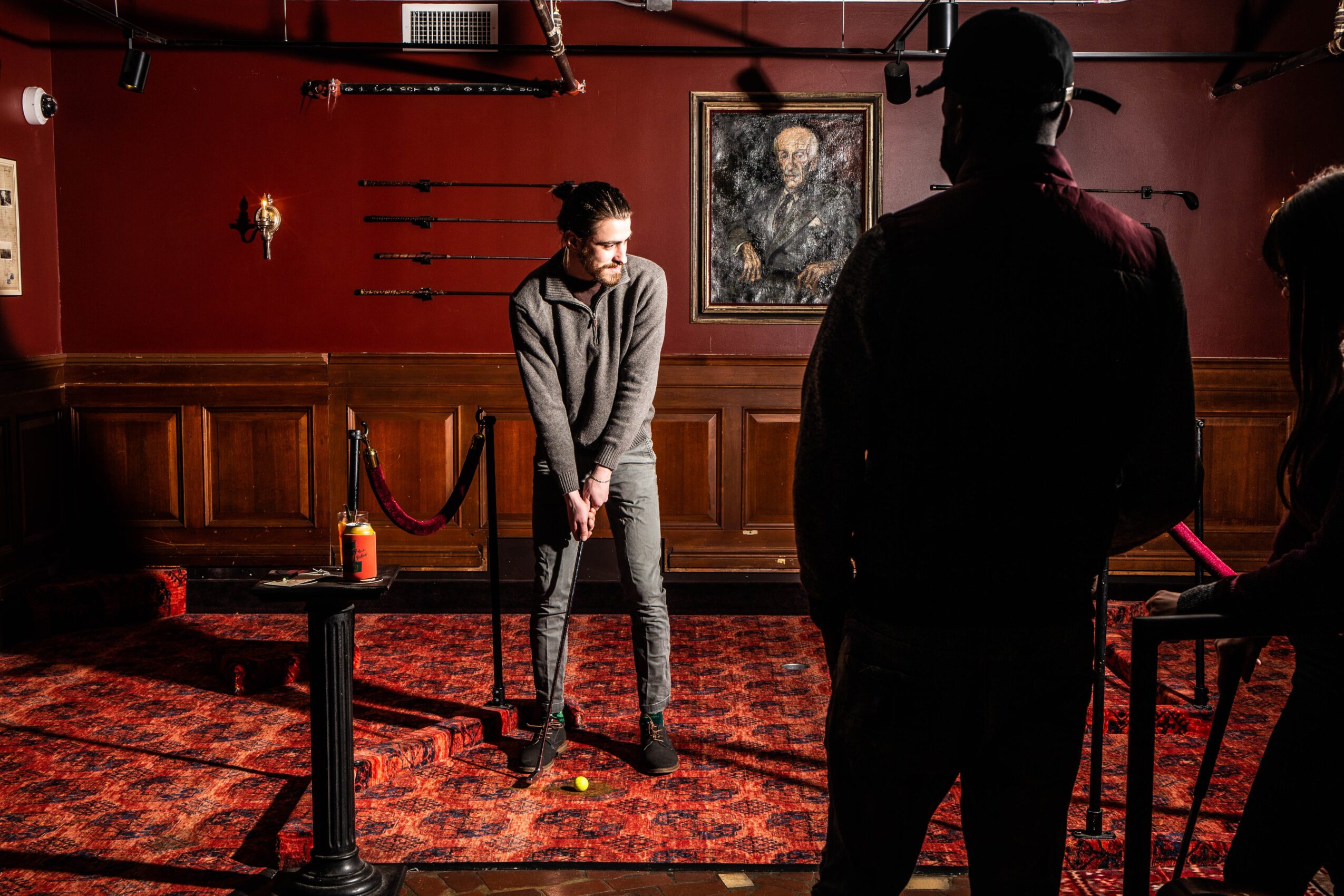 Person standing in a room with red walls, wooden paneling, and artwork, holding a golf club, with others nearby.