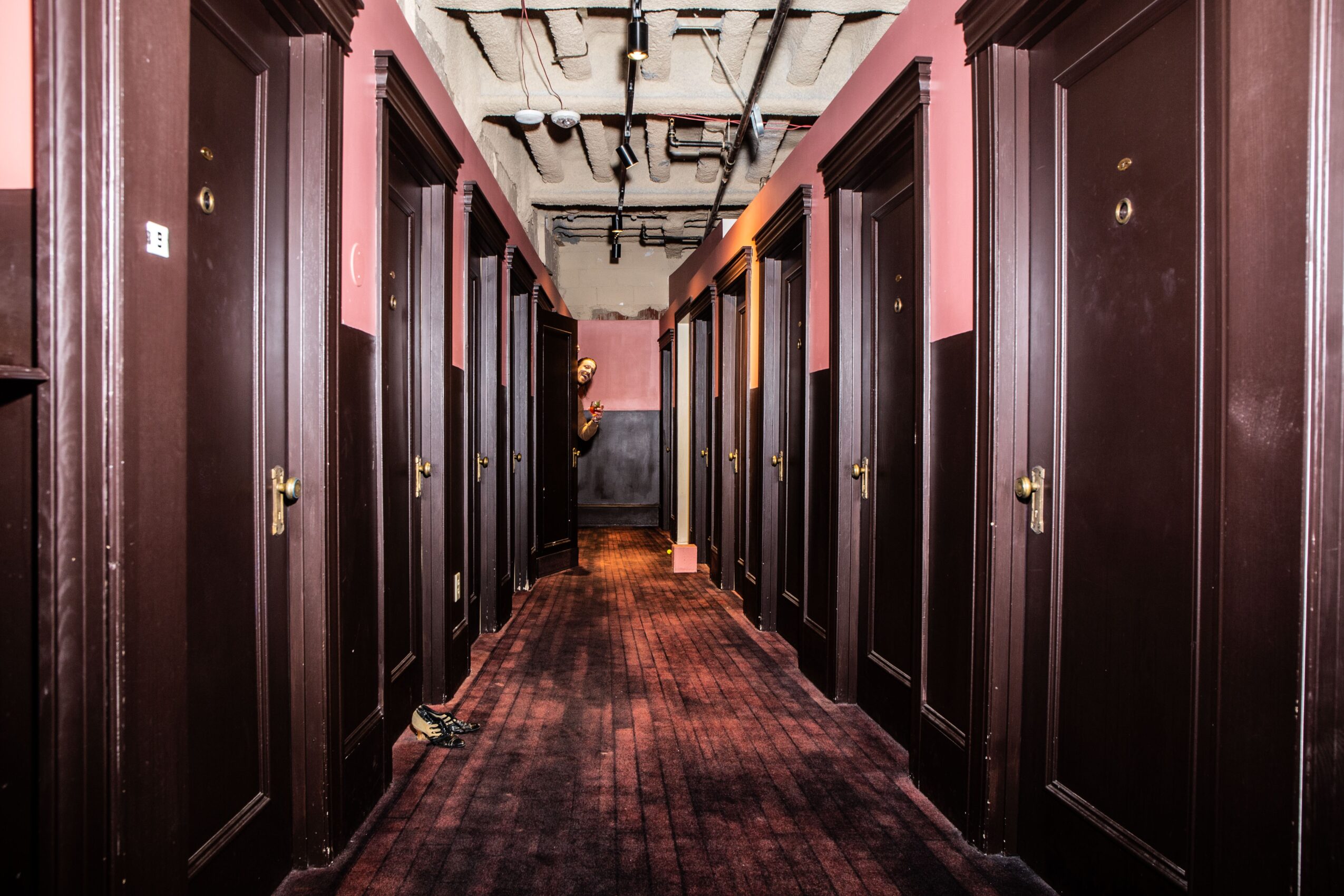 Long corridor with dark wooden doors on both sides, carpeted floor, and exposed ceiling pipes, leading to a distant wall.
