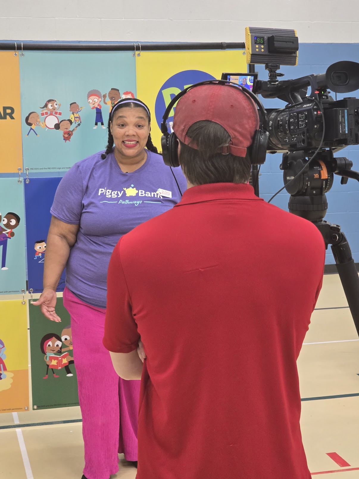 Person in red shirt filming woman in purple shirt with colorful background, including cartoon characters and a yellow sign.