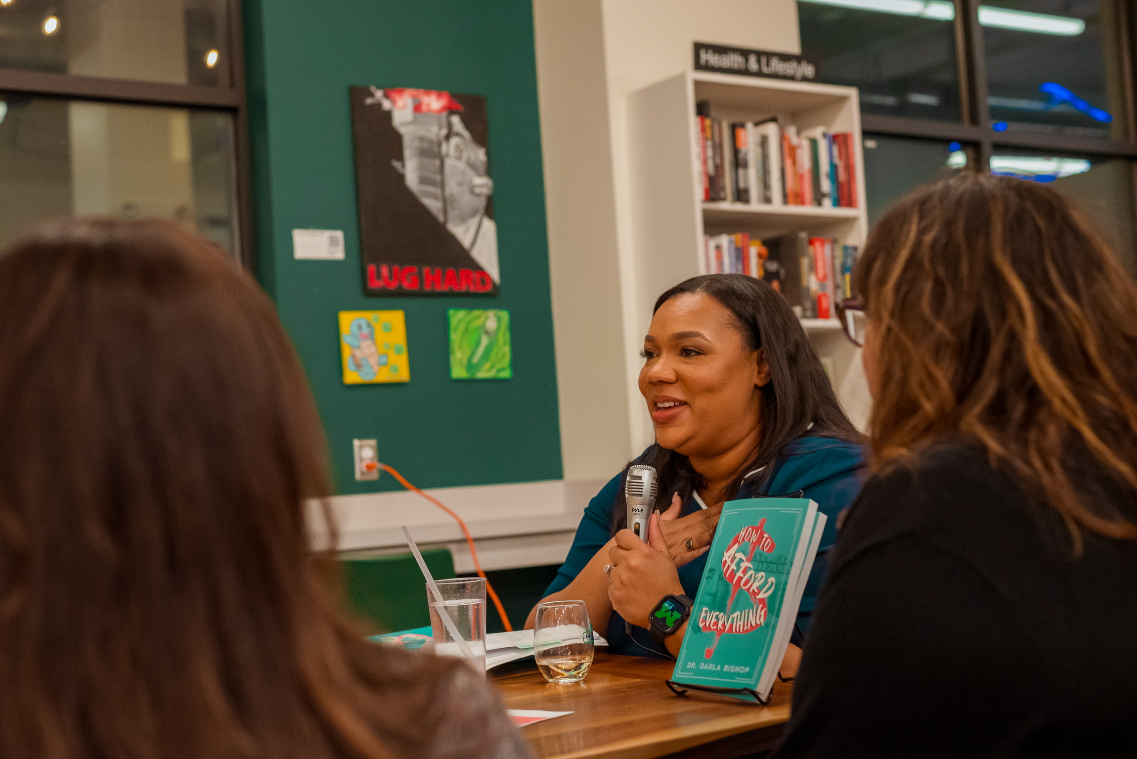 Three women sitting at a table in a library, engaged in conversation, with bookshelves in the background.