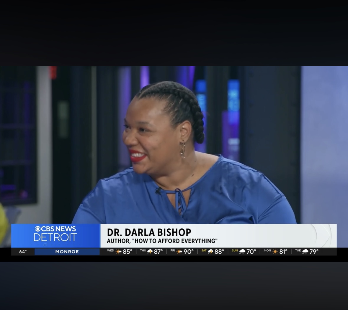 Woman with braided hair smiling, wearing a blue top, on a TV news set with a banner showing her name and book title.