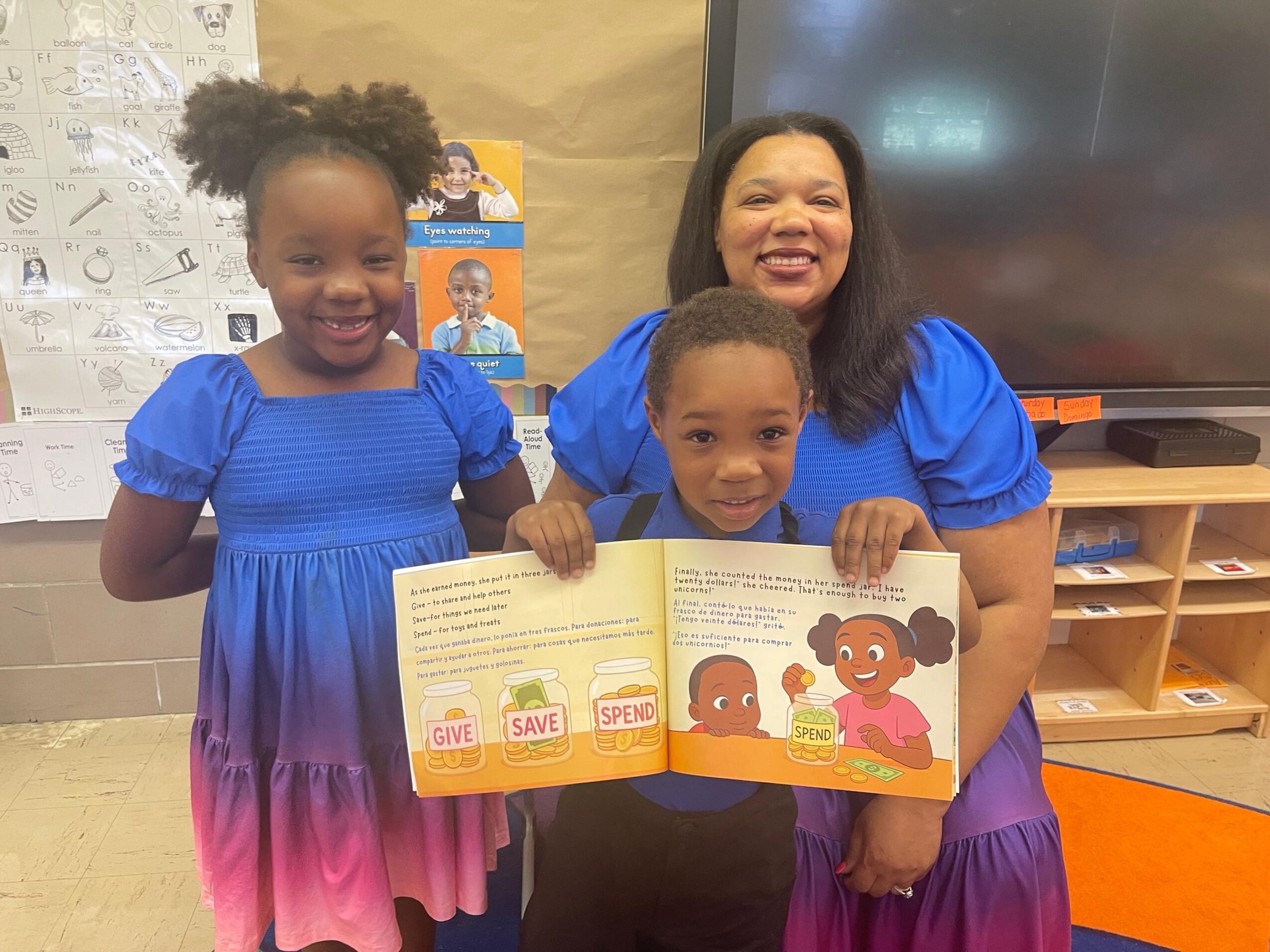 Smiling woman with two children holding a colorful book in a classroom, all wearing blue dresses.