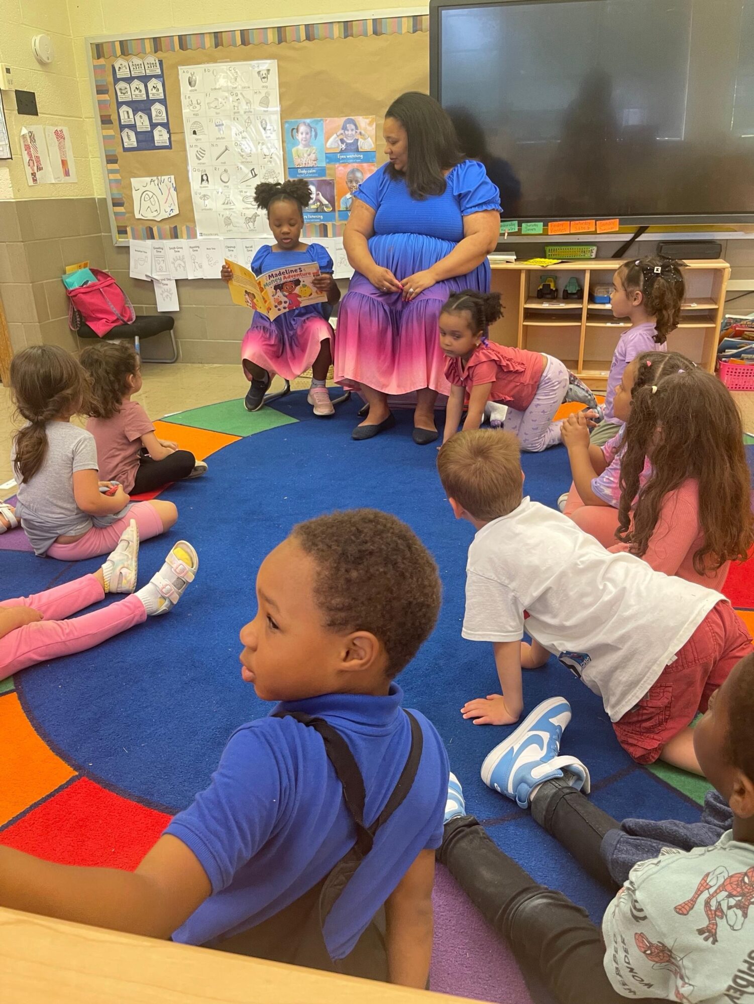 Teacher with children sitting on a colorful carpet in a classroom, some reading and others listening attentively.
