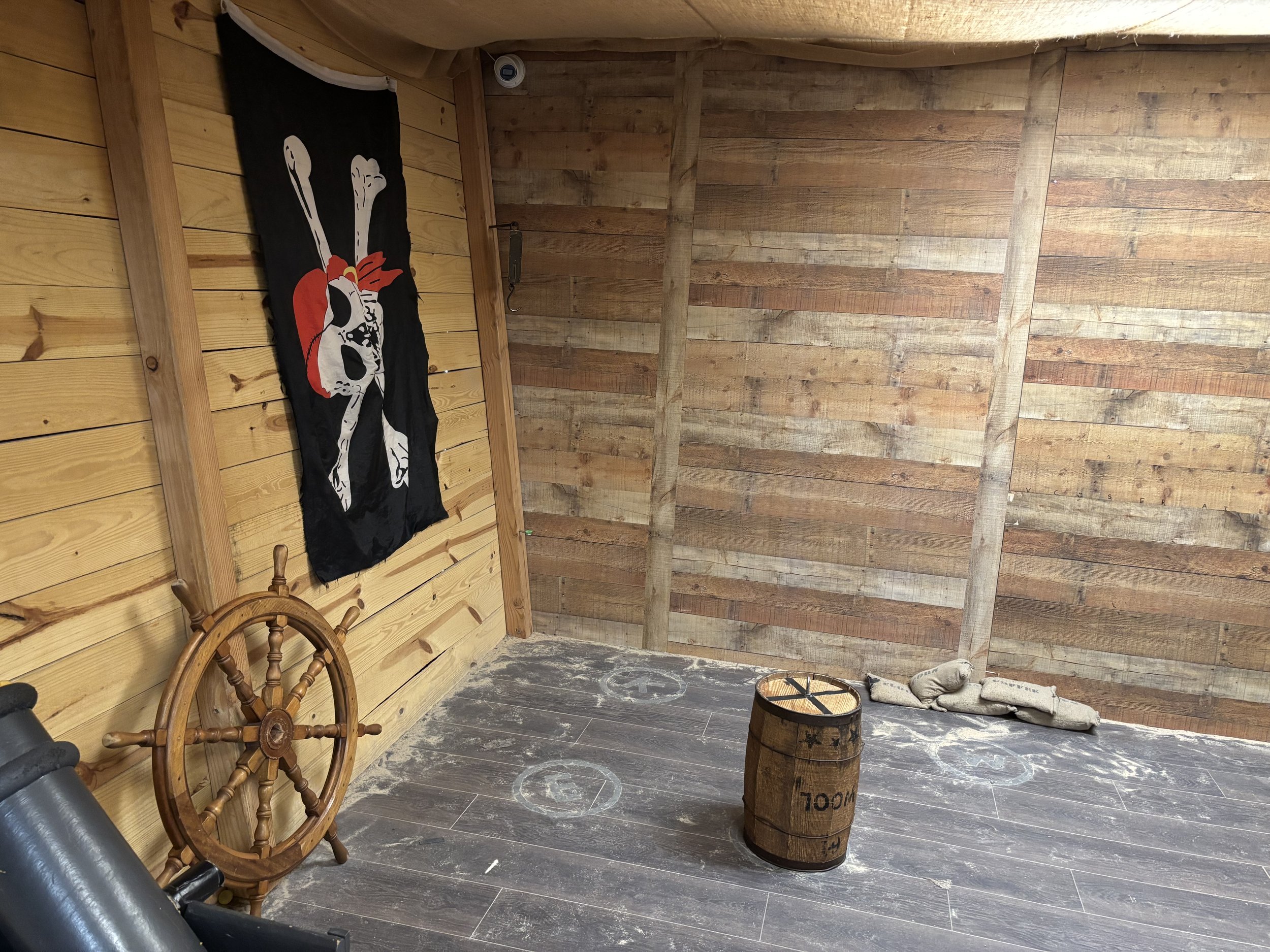 Wooden barrel with markings, ship's wheel, and a skull and crossbones flag inside a wooden room.