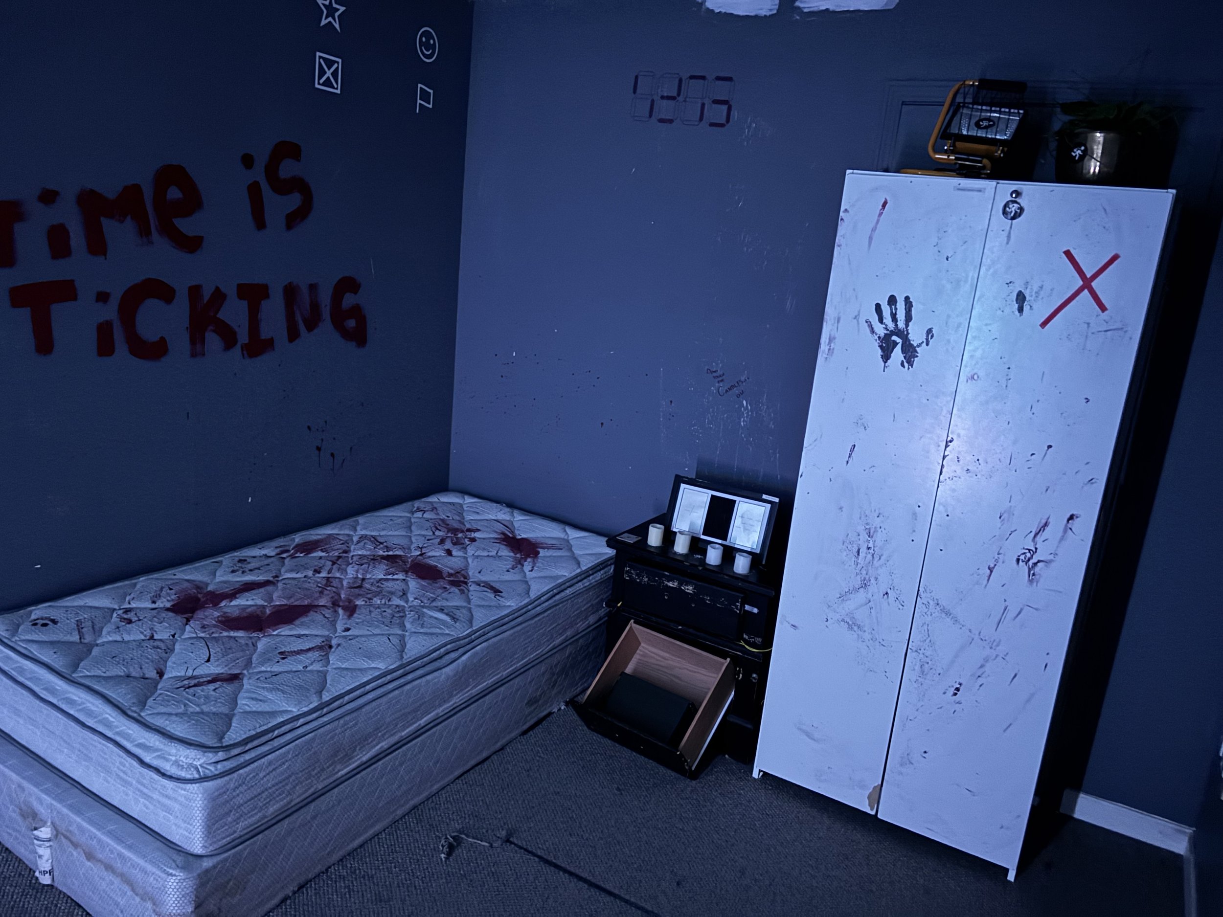 A room with a mattress, white wardrobe with handprints, and a wall with graffiti. The room appears dark and cluttered.