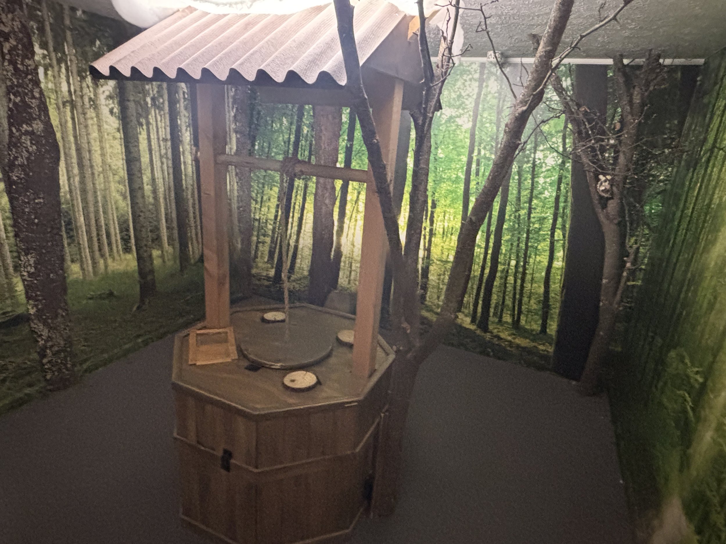 Wooden well with a roof inside a forest-themed room, with trees and greenery on the walls.