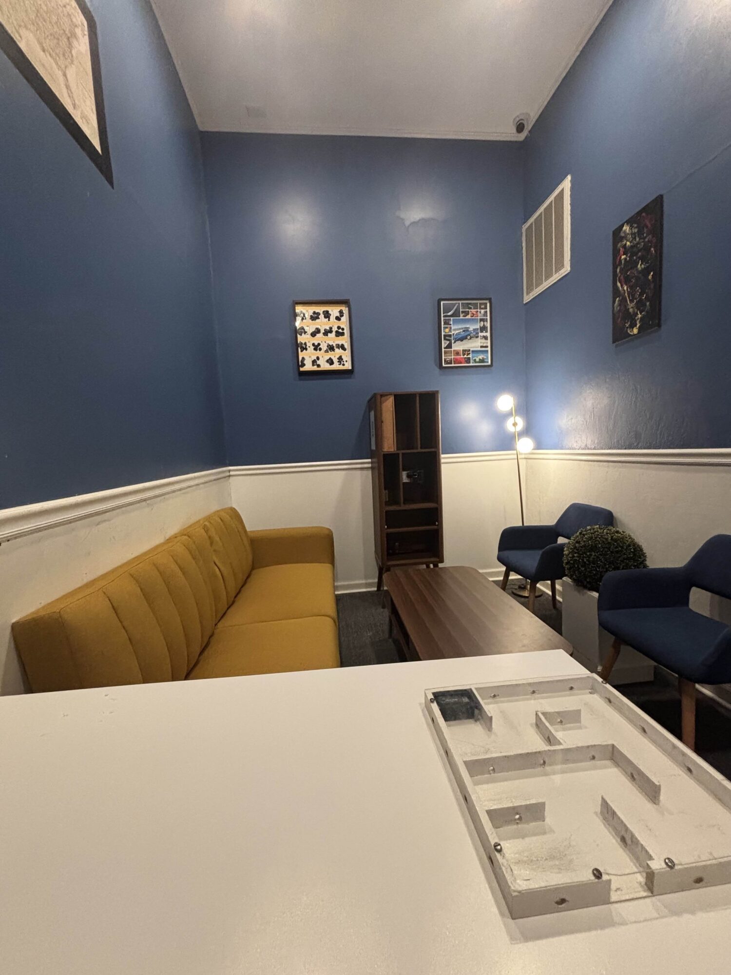 Room with blue walls, yellow sofa, two blue chairs, and artwork on walls, viewed from a table with a clear plastic container.
