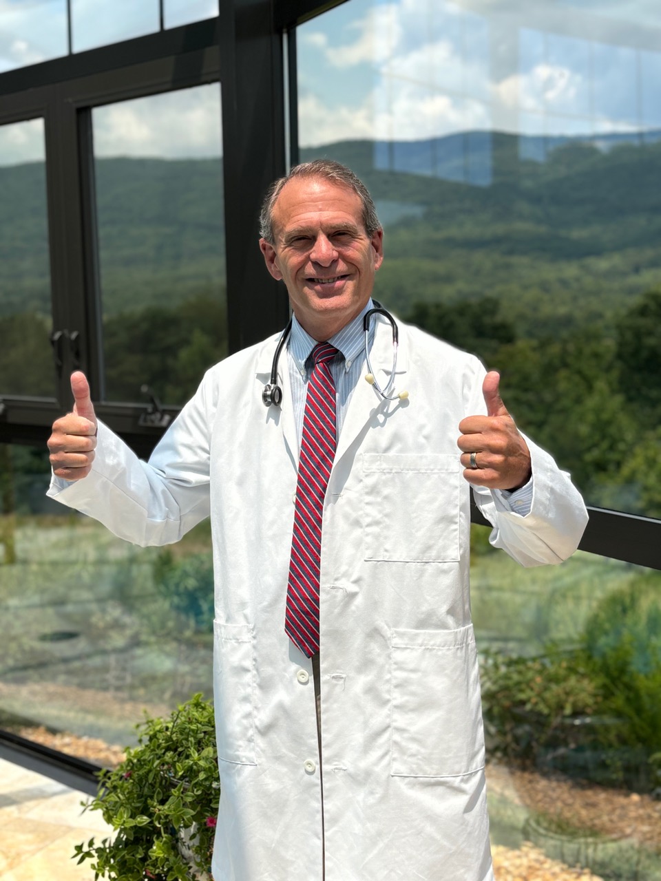 Doctor in white coat with stethoscope giving two thumbs up, standing indoors with large window and outdoor landscape behind.