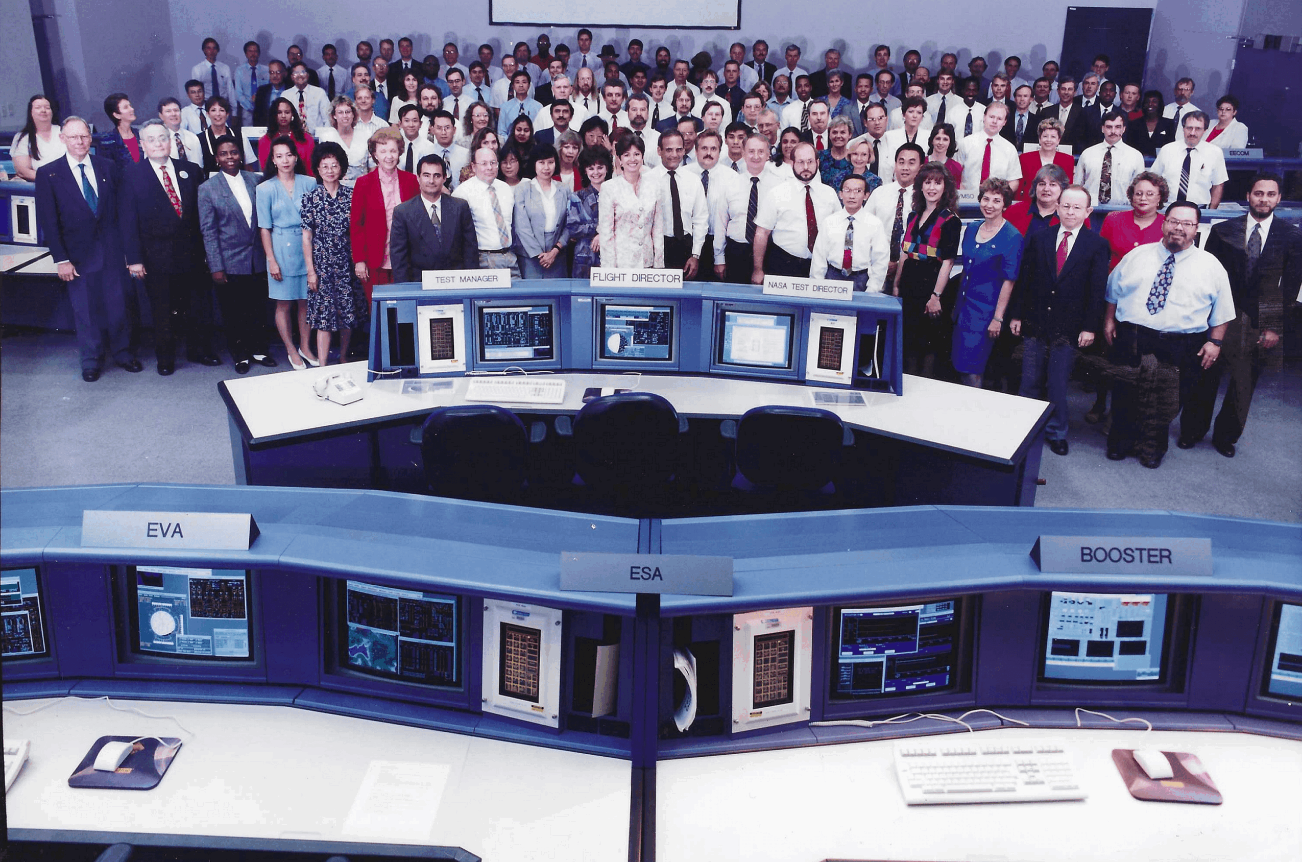 Large group of people in a control room with multiple computer monitors and workstations.