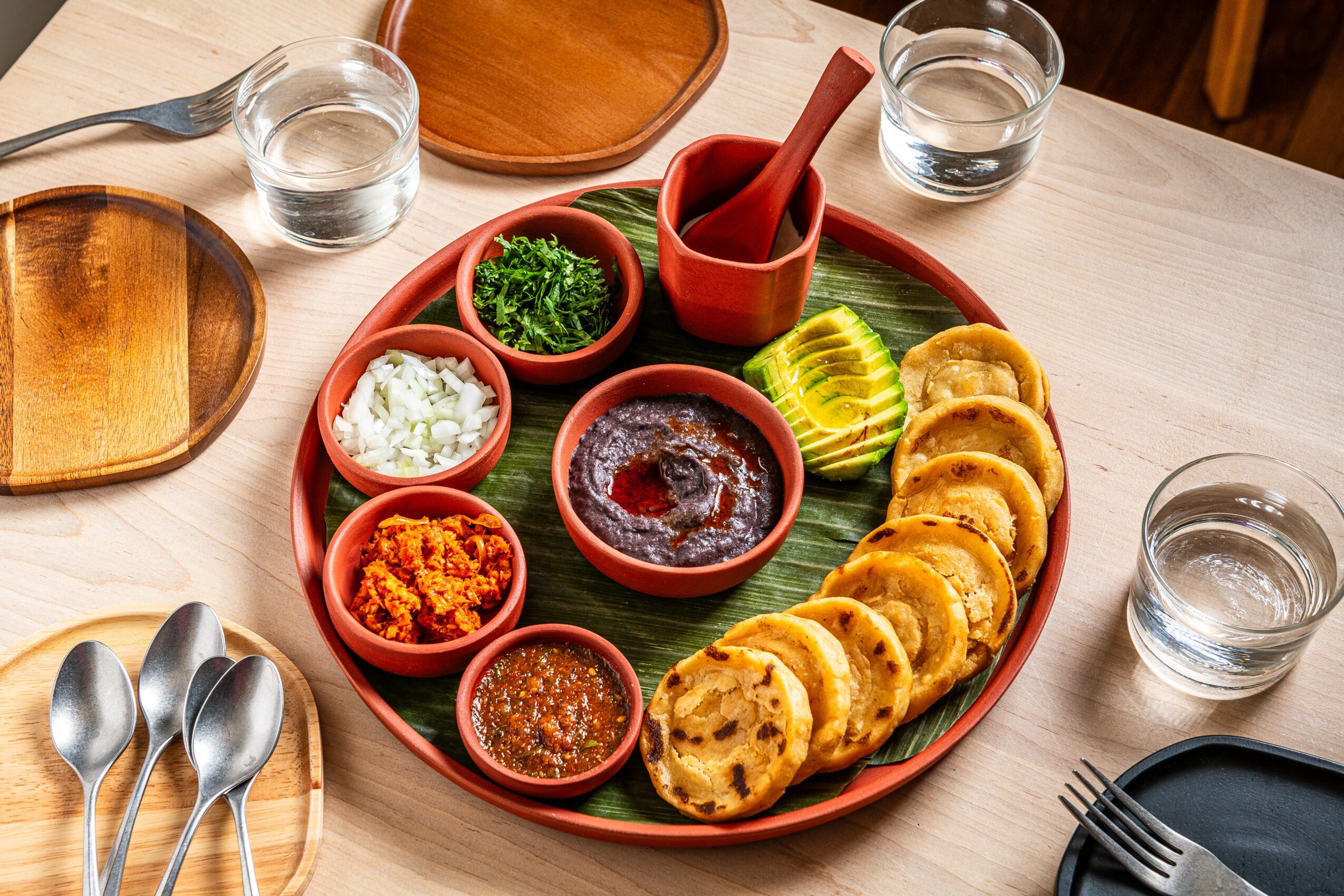 Plate with various Indian snacks, chutneys, and dips, surrounded by glasses of water and empty plates on a table.